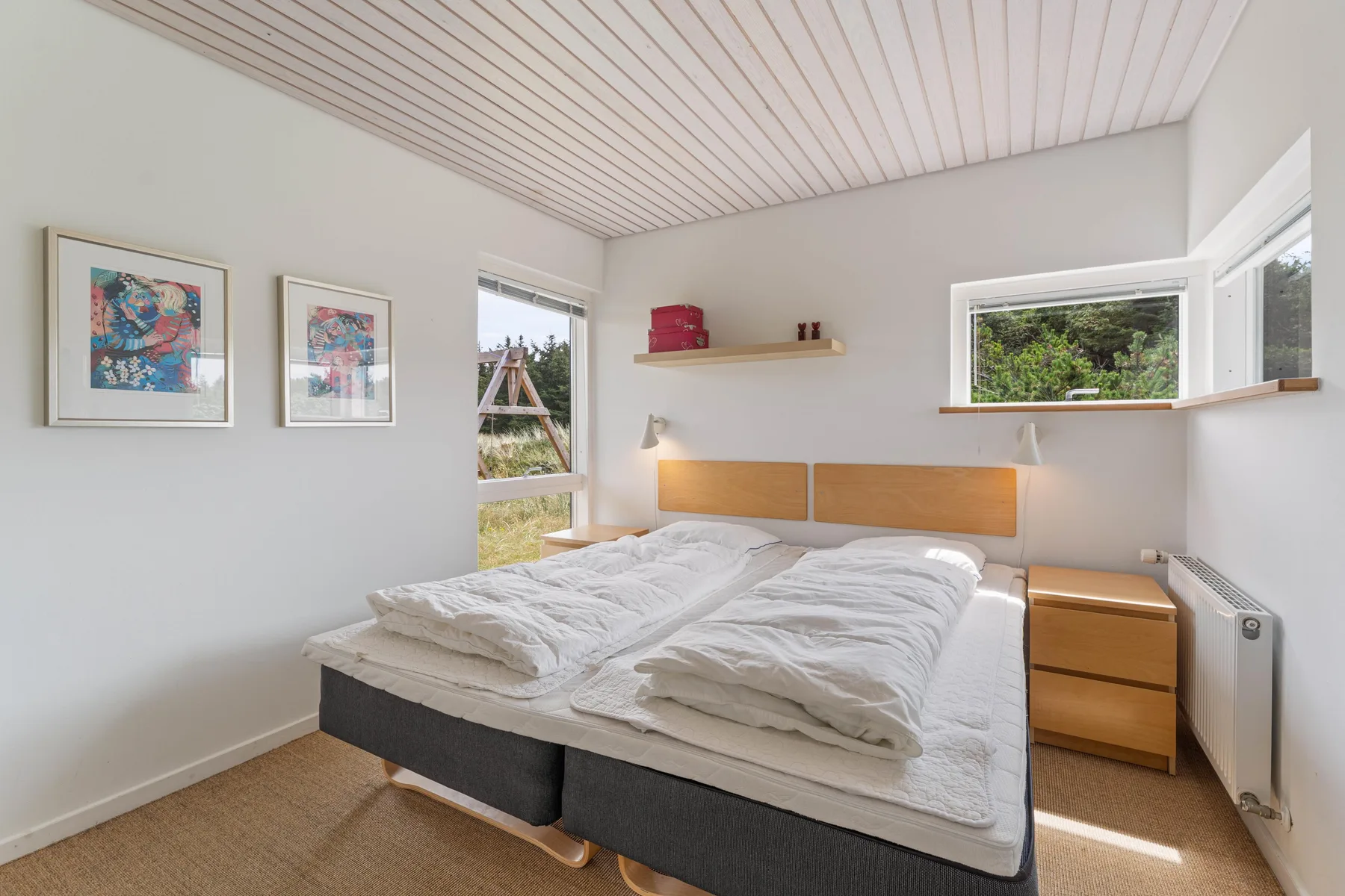 A bright bedroom with a double bed, framed artwork, and windows.