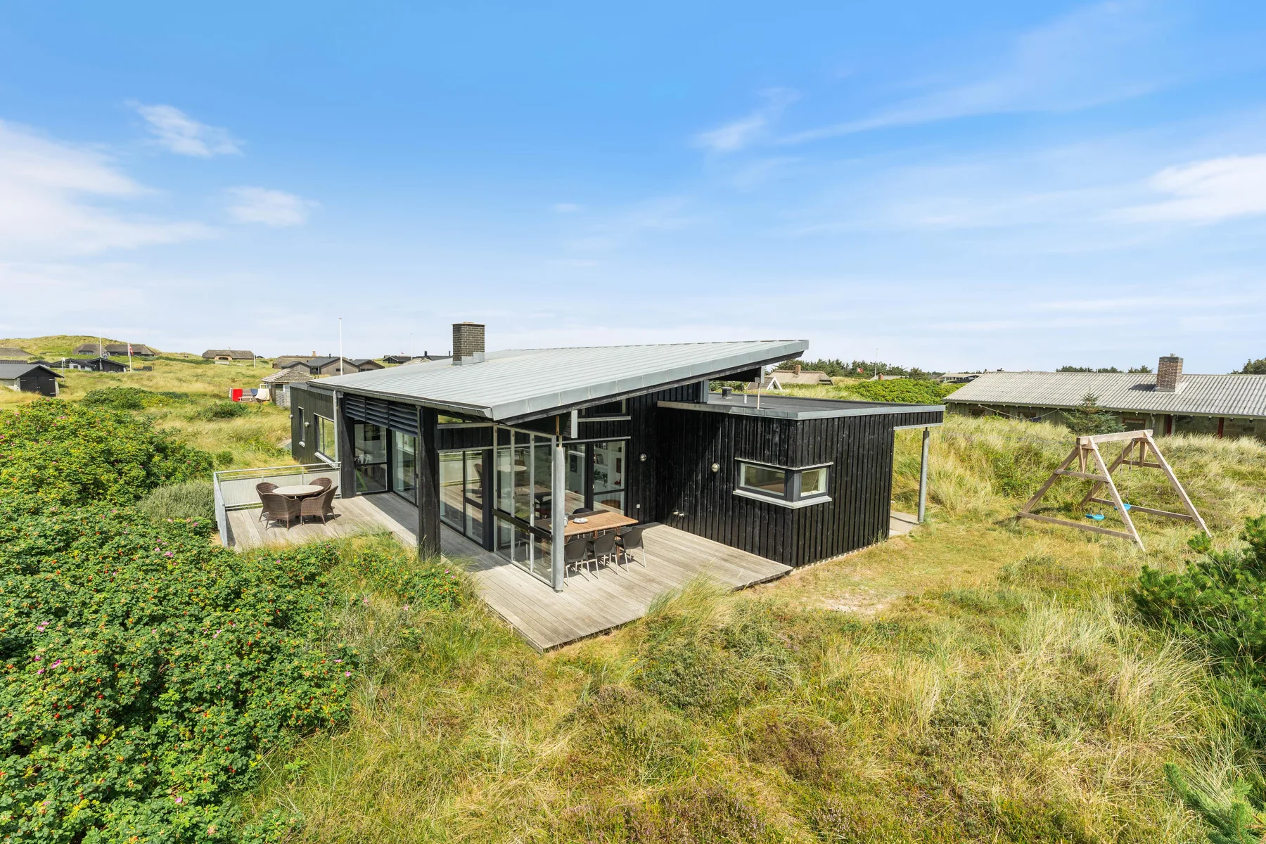 A modern dark-clad house sits among grassy dunes beneath a blue sky.