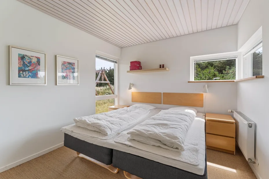 A bright bedroom with two beds, framed artwork, and windows overlooking greenery.