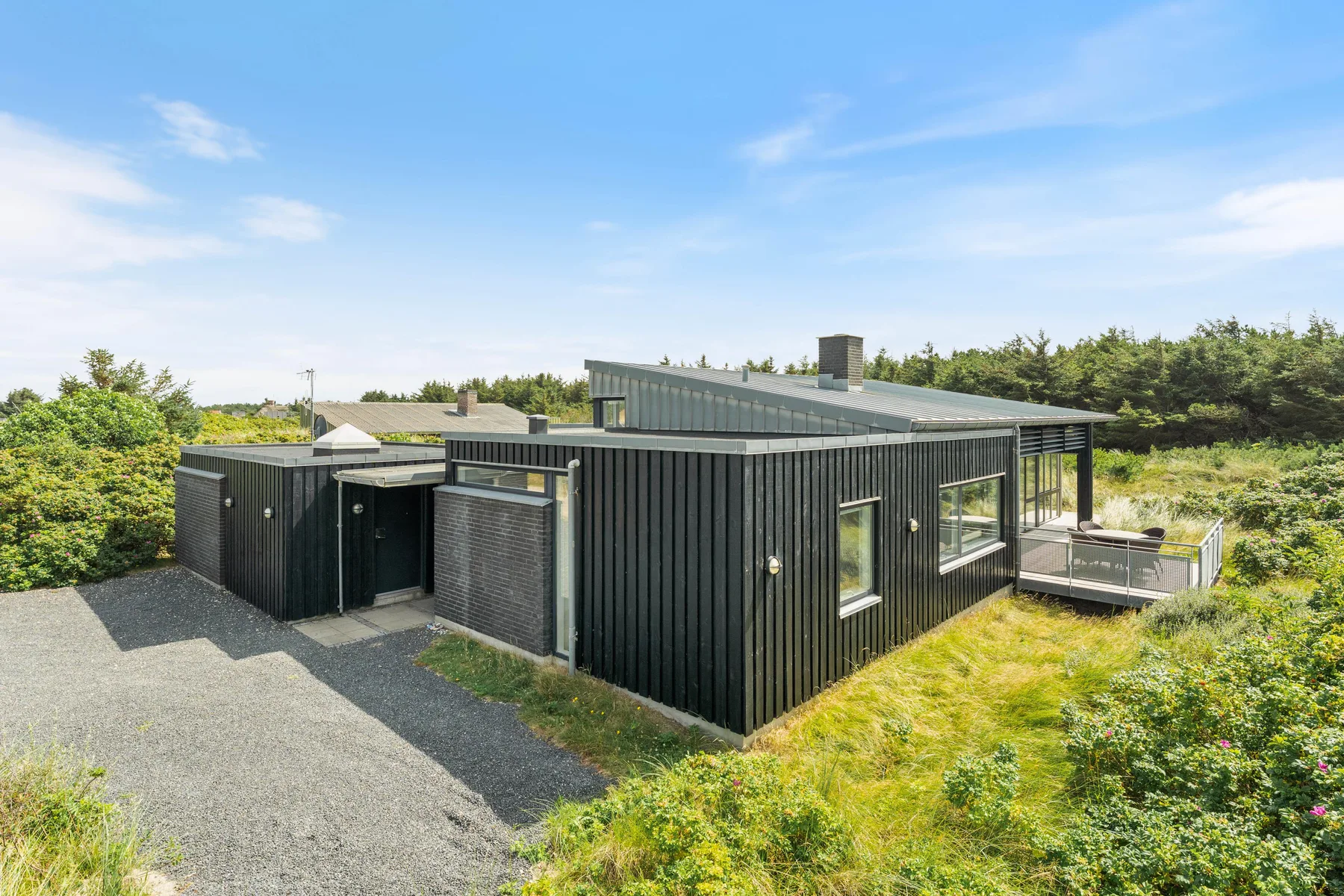 A modern dark-sided house with a gravel driveway and surrounding greenery.