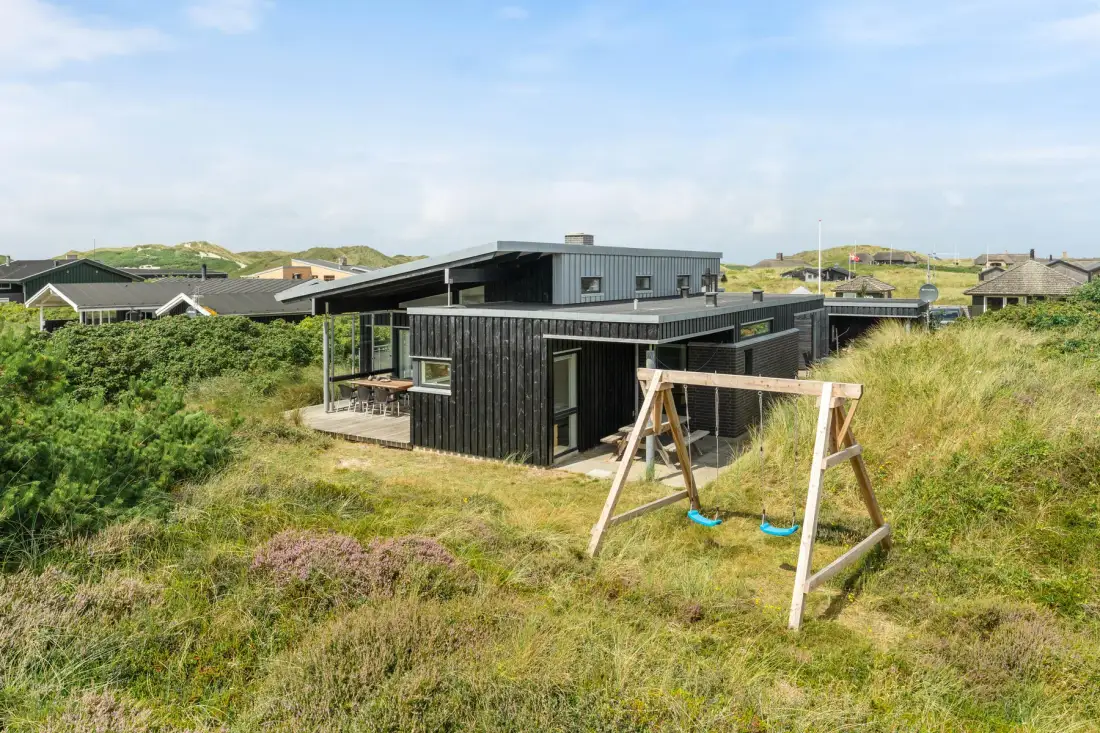 A modern house sits among grassy dunes, with a wooden swing set in the foreground.