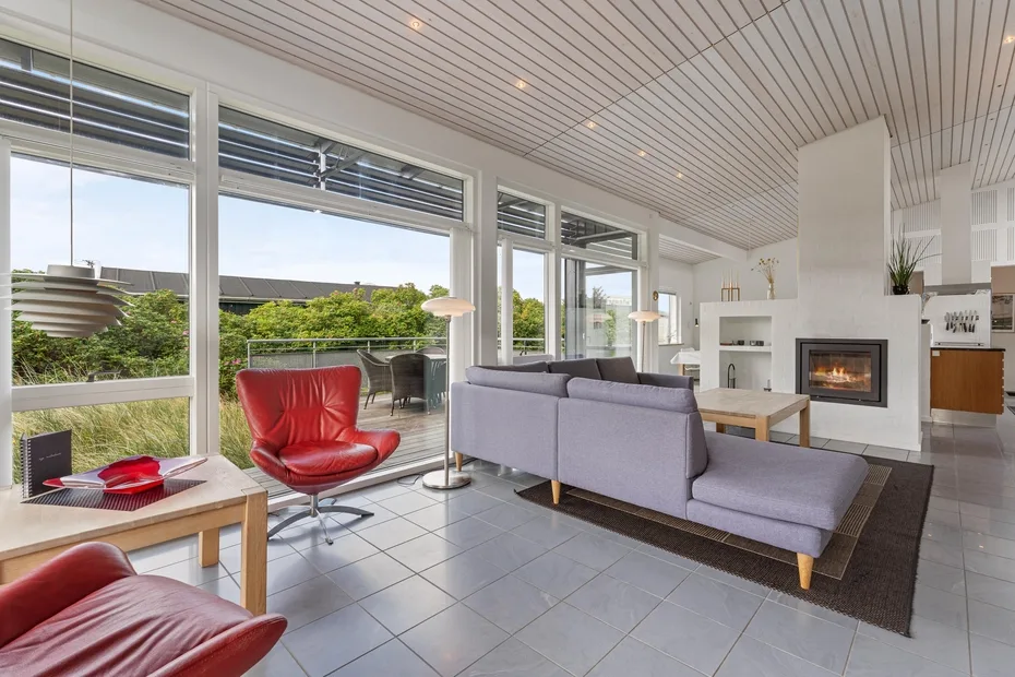 A bright living room with a gray sofa, red chair, and fireplace beside large windows.