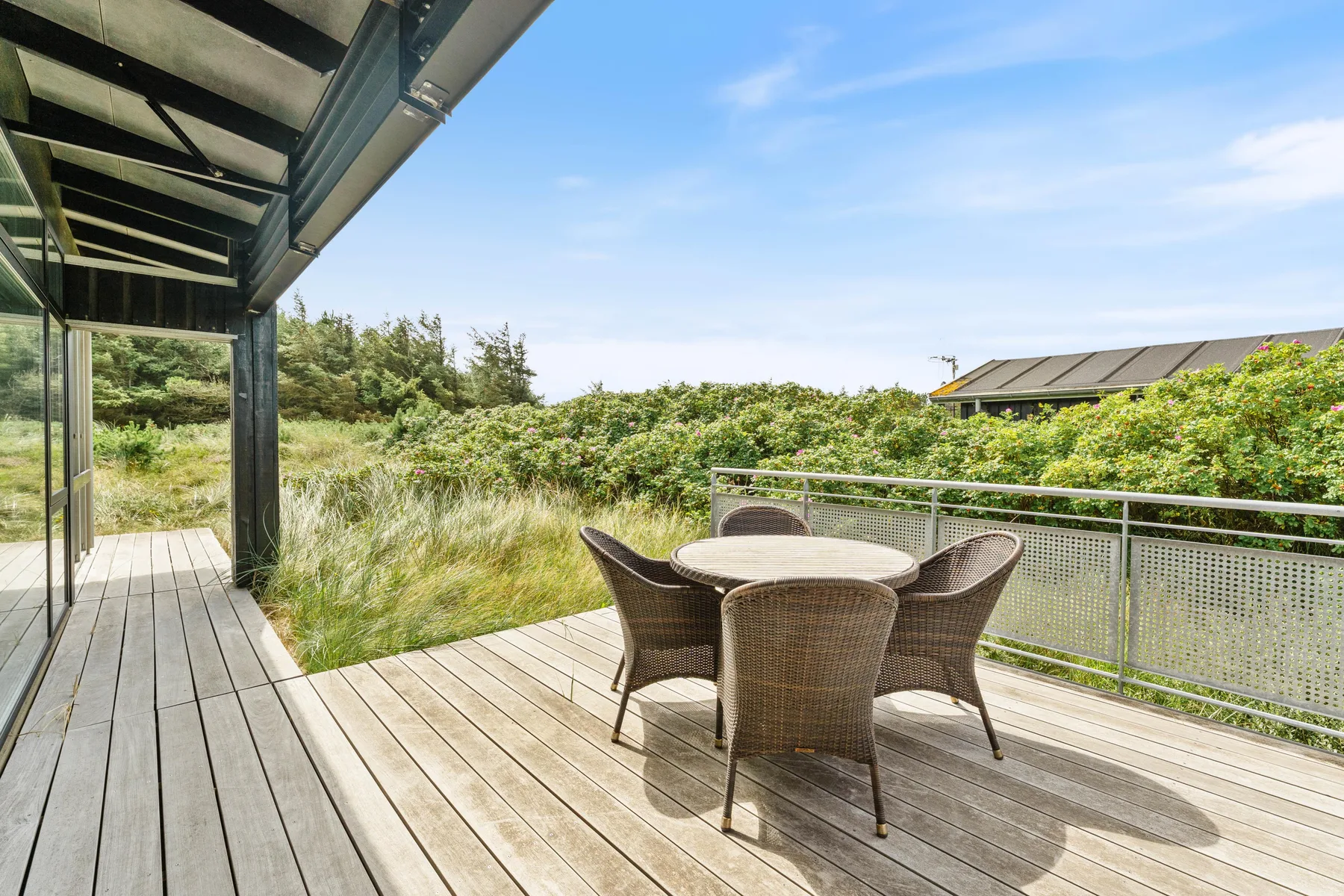 A wooden outdoor deck has a round table and seating beside green vegetation.