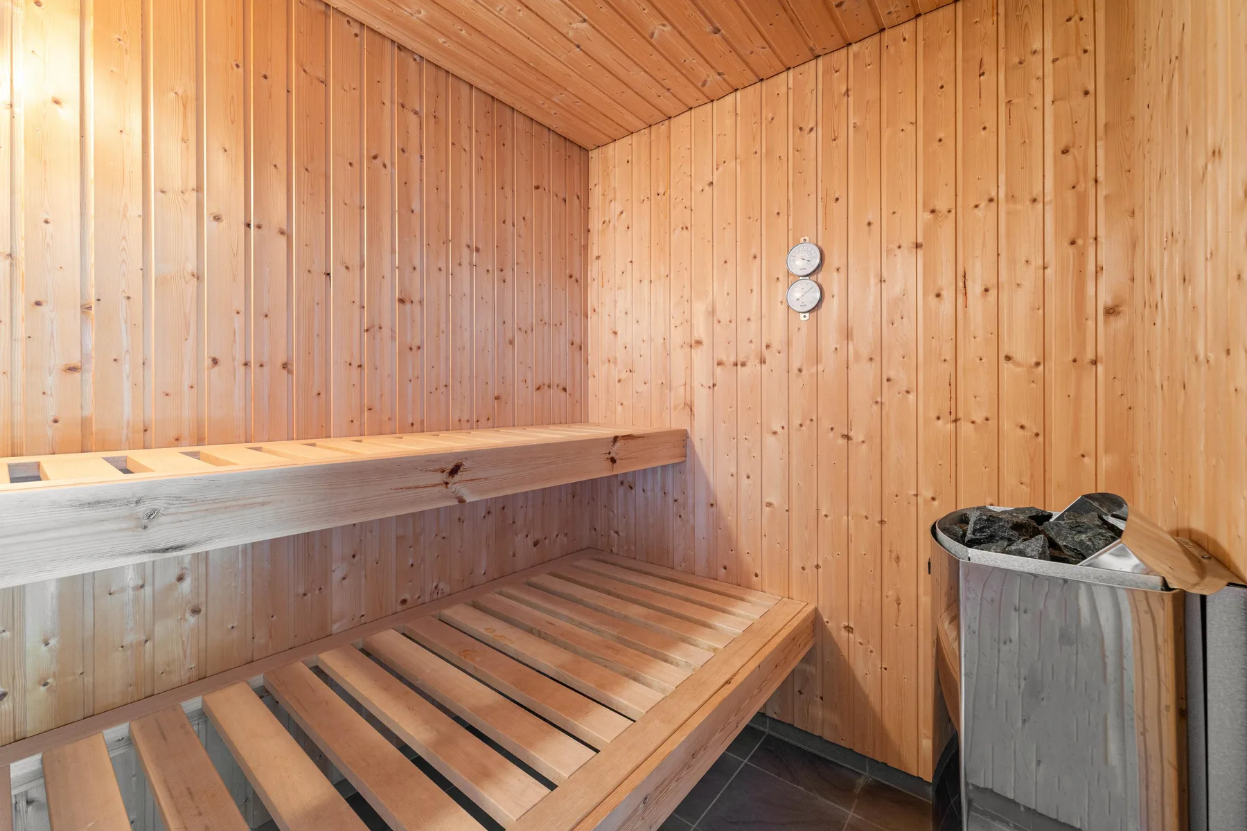 A wood-paneled sauna has tiered wooden seating and a heater filled with stones.