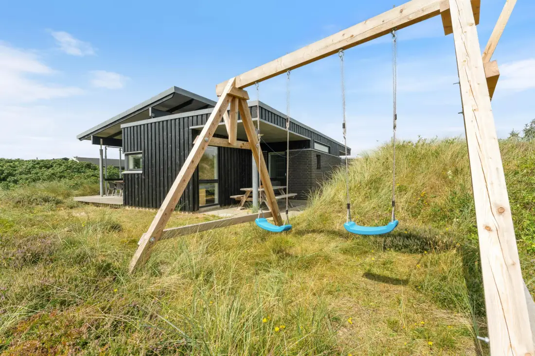 A dark modern house stands behind a wooden swing set in grassy dunes.