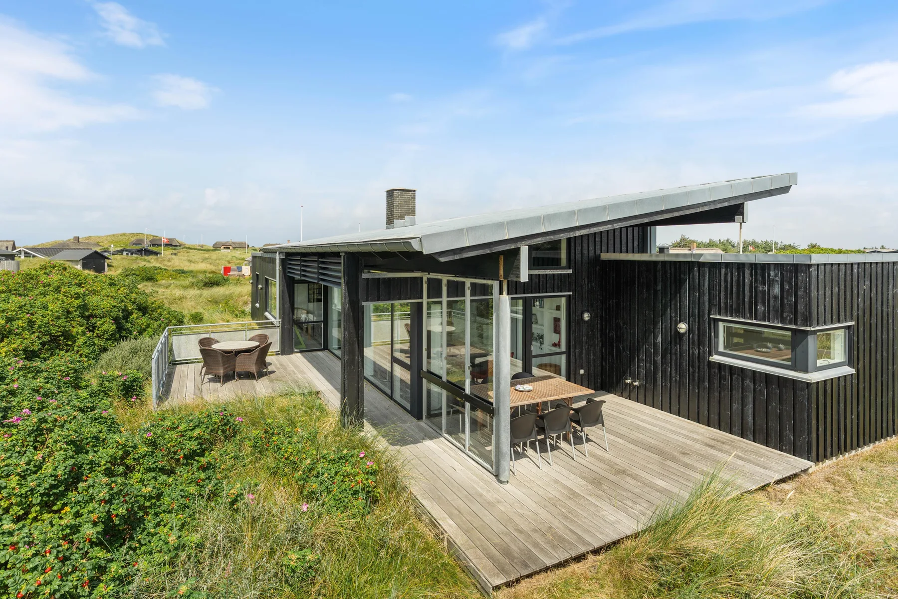 A modern dark-clad house with a broad wooden deck surrounded by greenery.