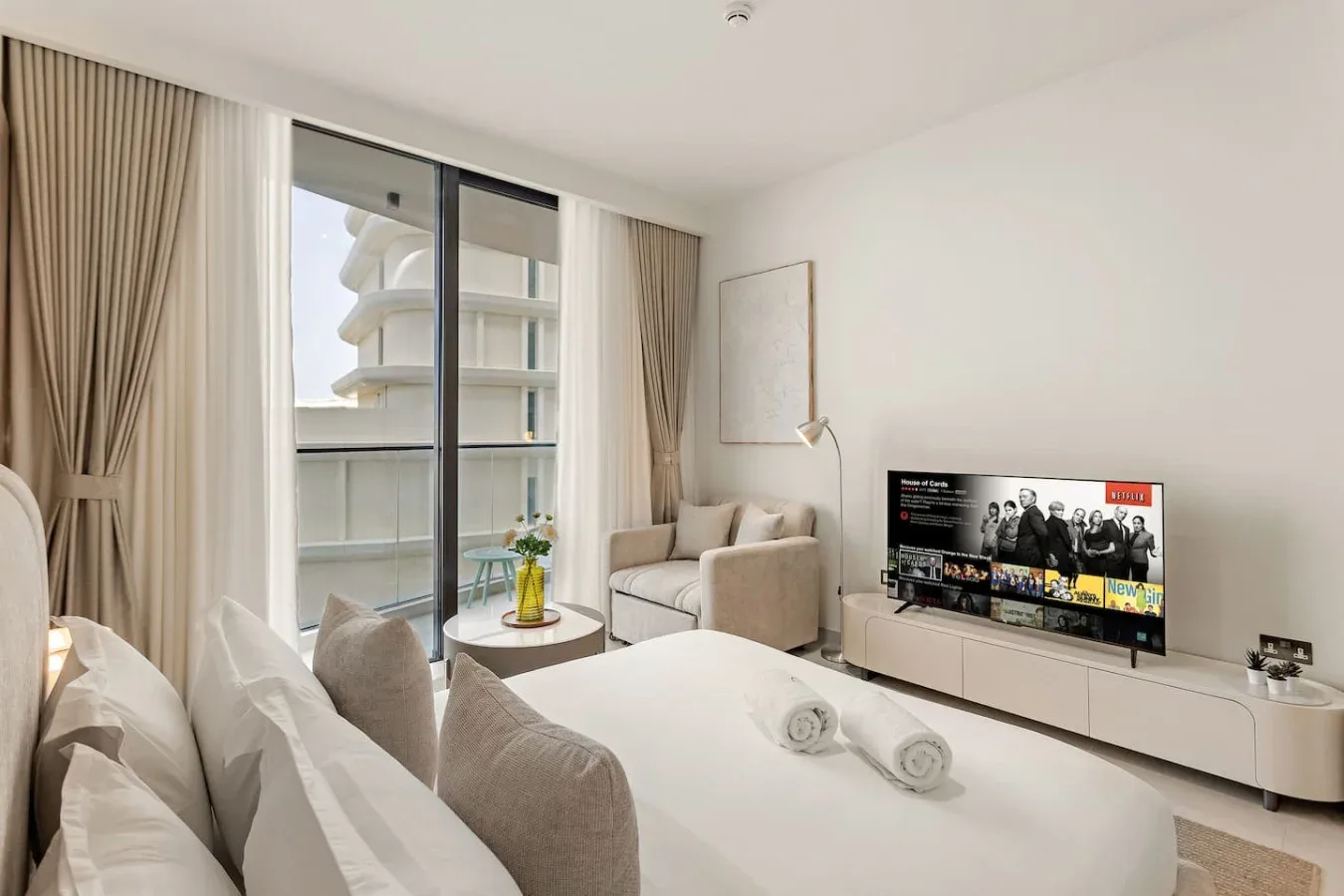 A bright bedroom with a bed, seating, and a television beside the window.