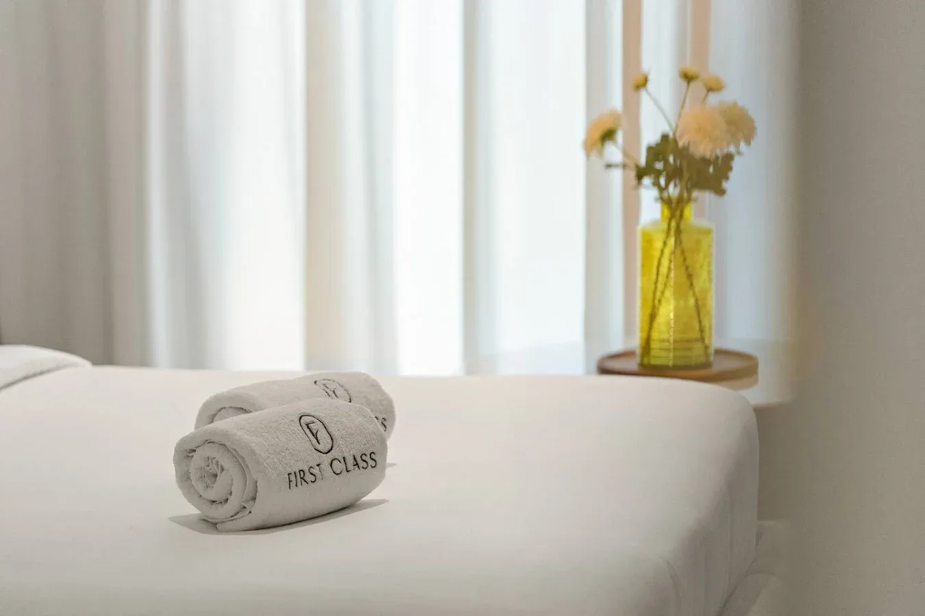 A neatly made bed with folded towels and a vase of flowers by the window.