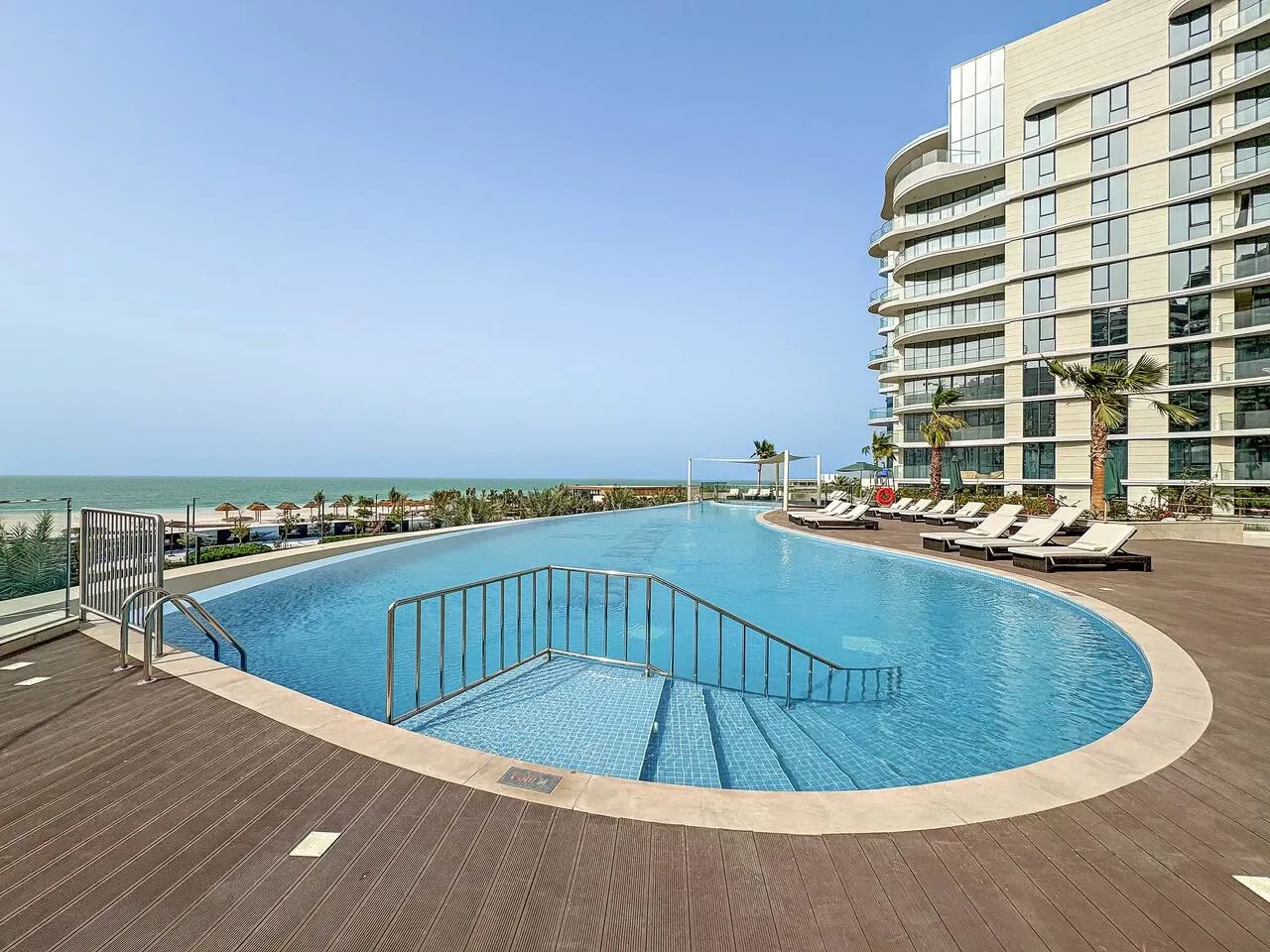 Una piscina exterior curva junto a un edificio moderno, con el mar visible a lo lejos.
