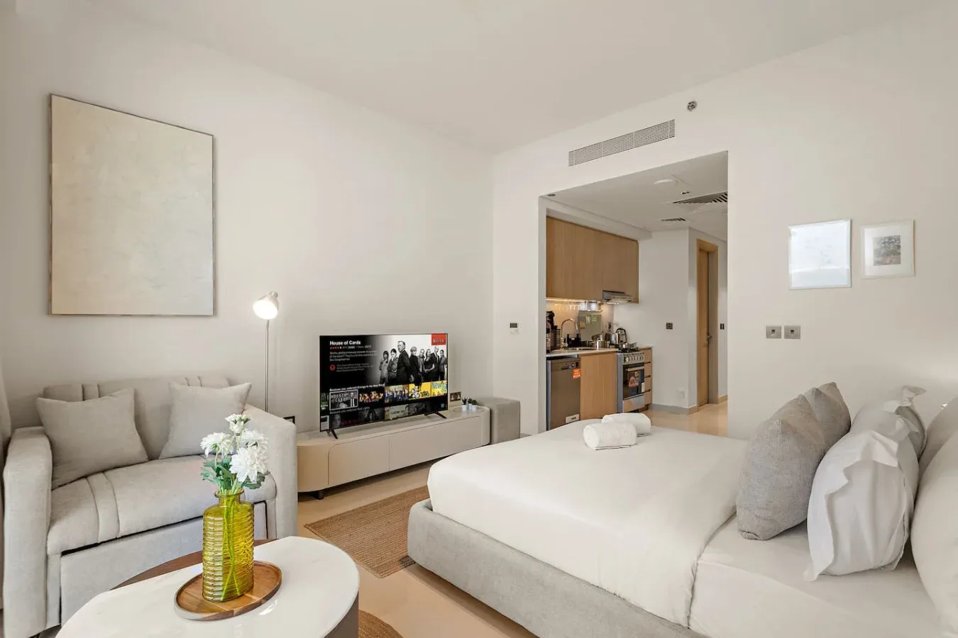 A bright bedroom with a large bed, light seating, and an open kitchenette.
