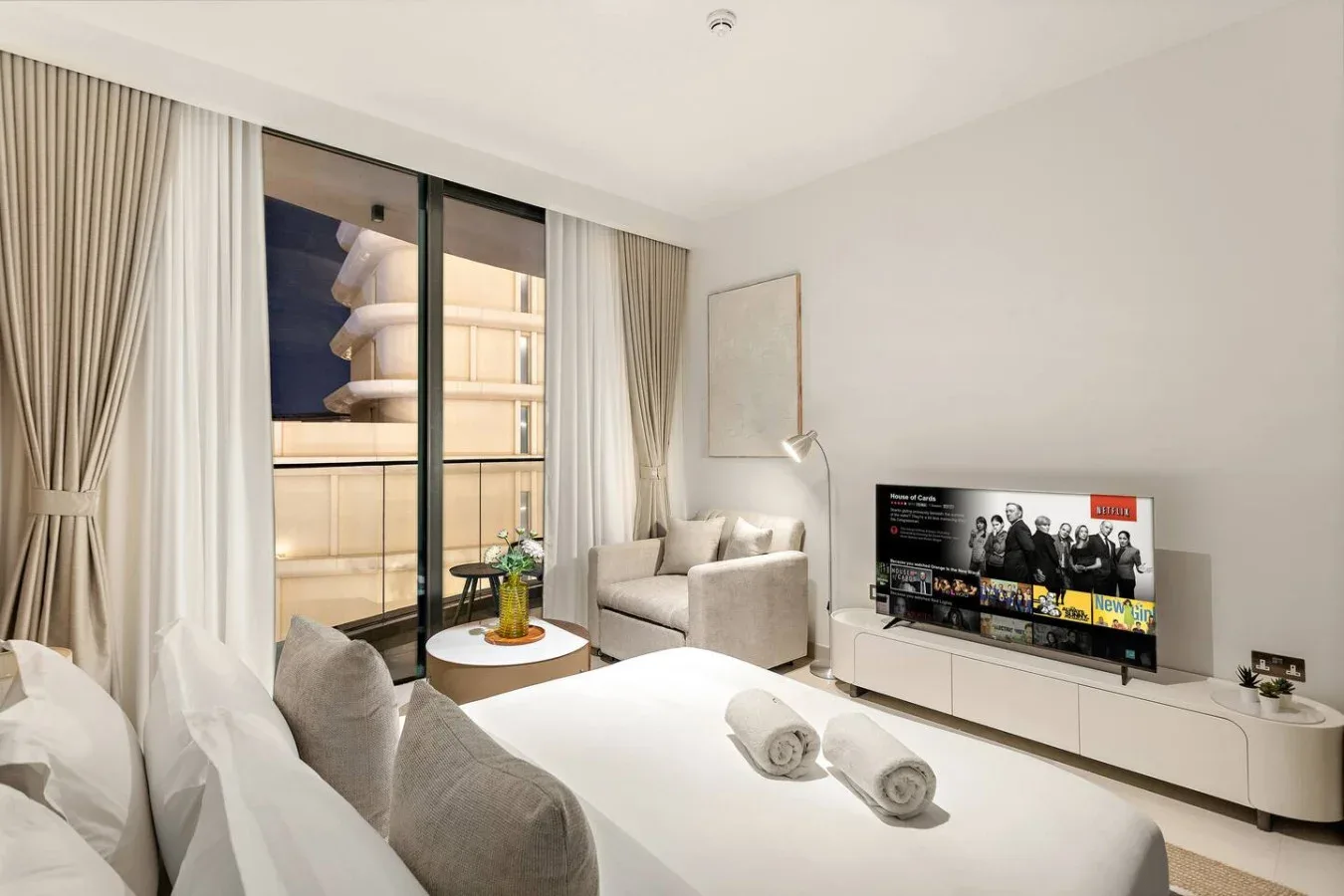 A bright bedroom with a bed, seating area, and television beside glass doors.