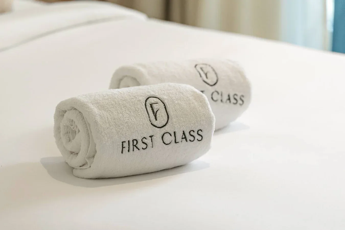 Two rolled towels embroidered with “First Class” rest on a neatly made bed.