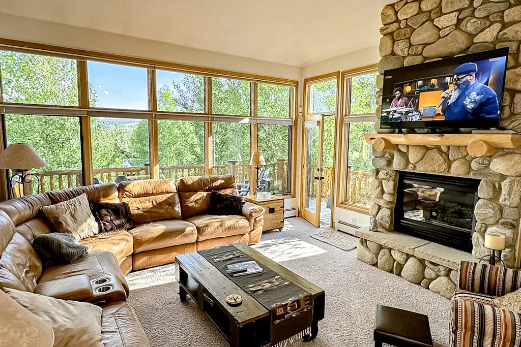 A living room with large windows, seating, and a stone fireplace.