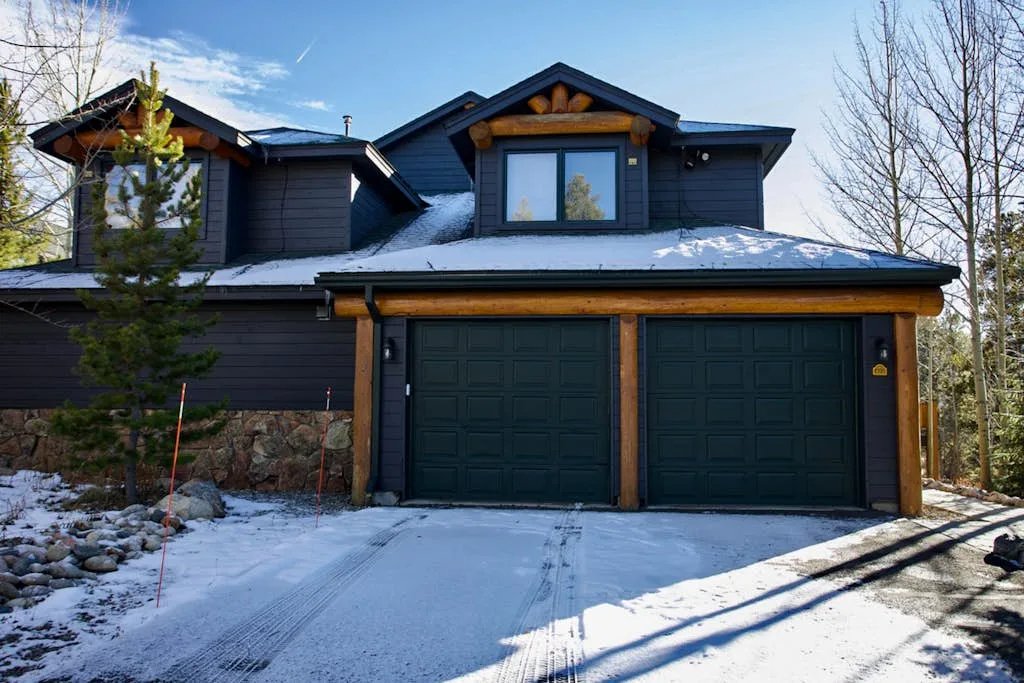 A dark-sided house with two garage doors stands beside a snowy driveway.