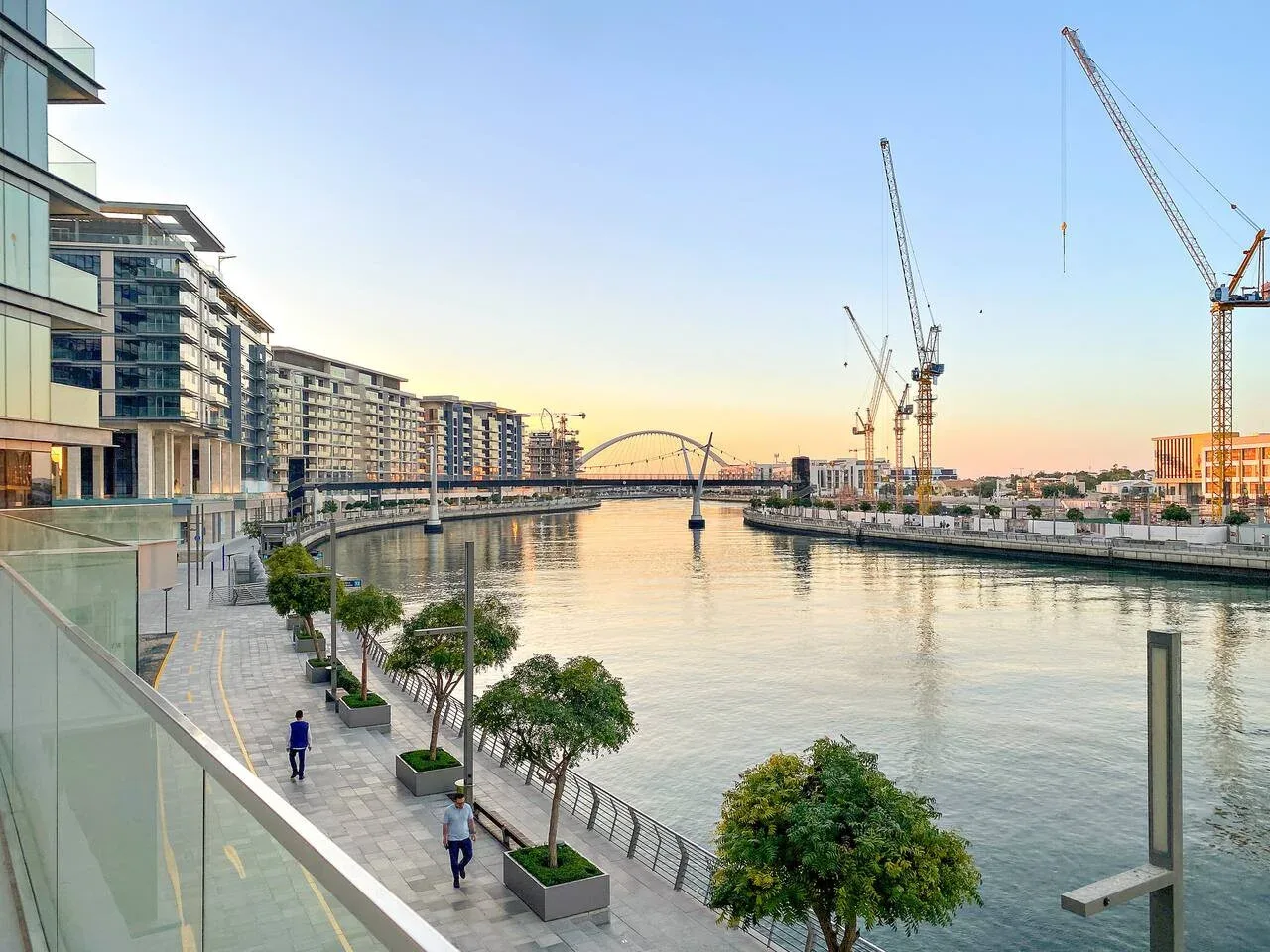 A waterfront promenade borders a calm canal, with buildings and construction cranes nearby.