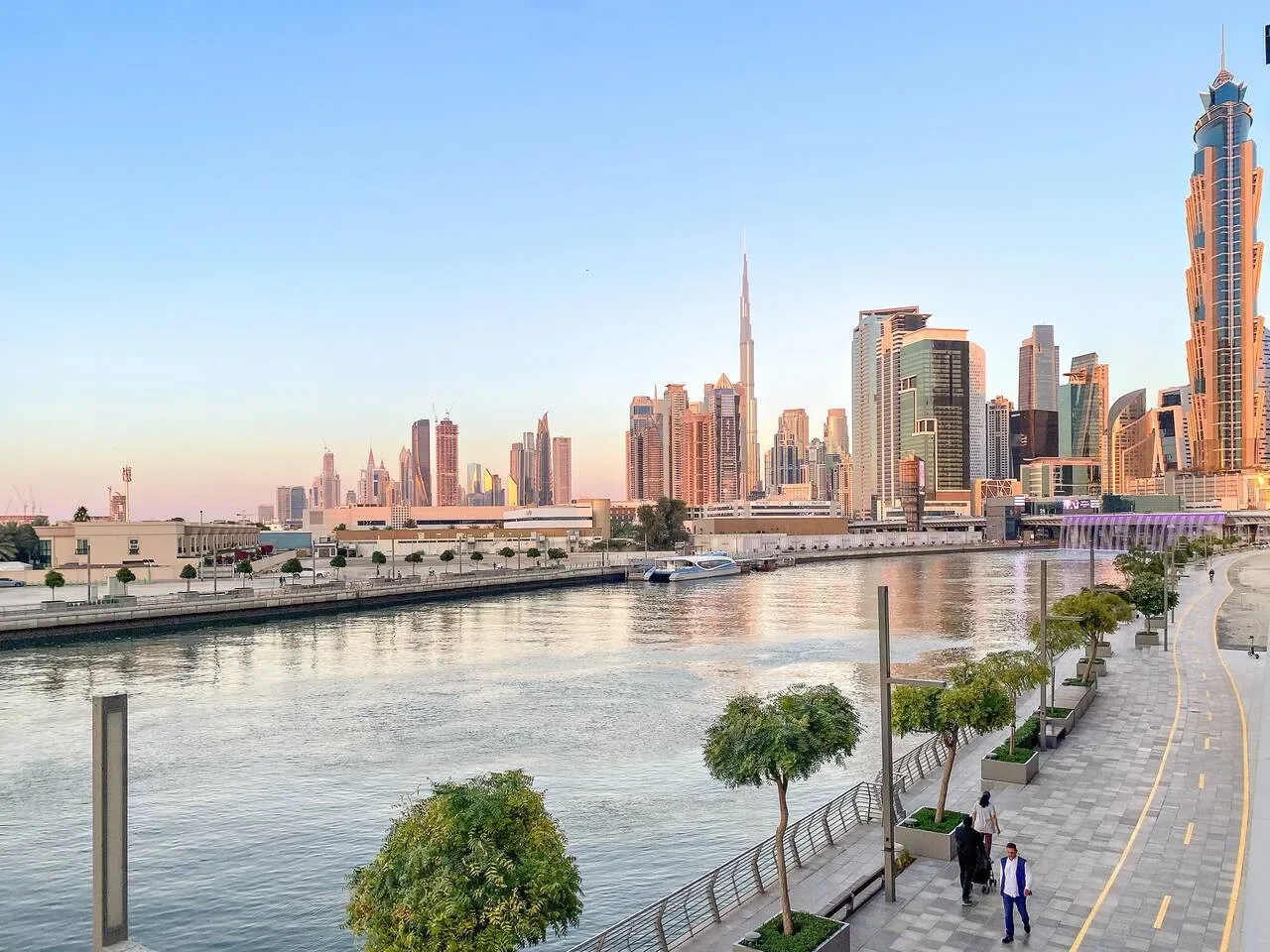 A waterfront promenade runs beside a canal, with a city skyline in the background.
