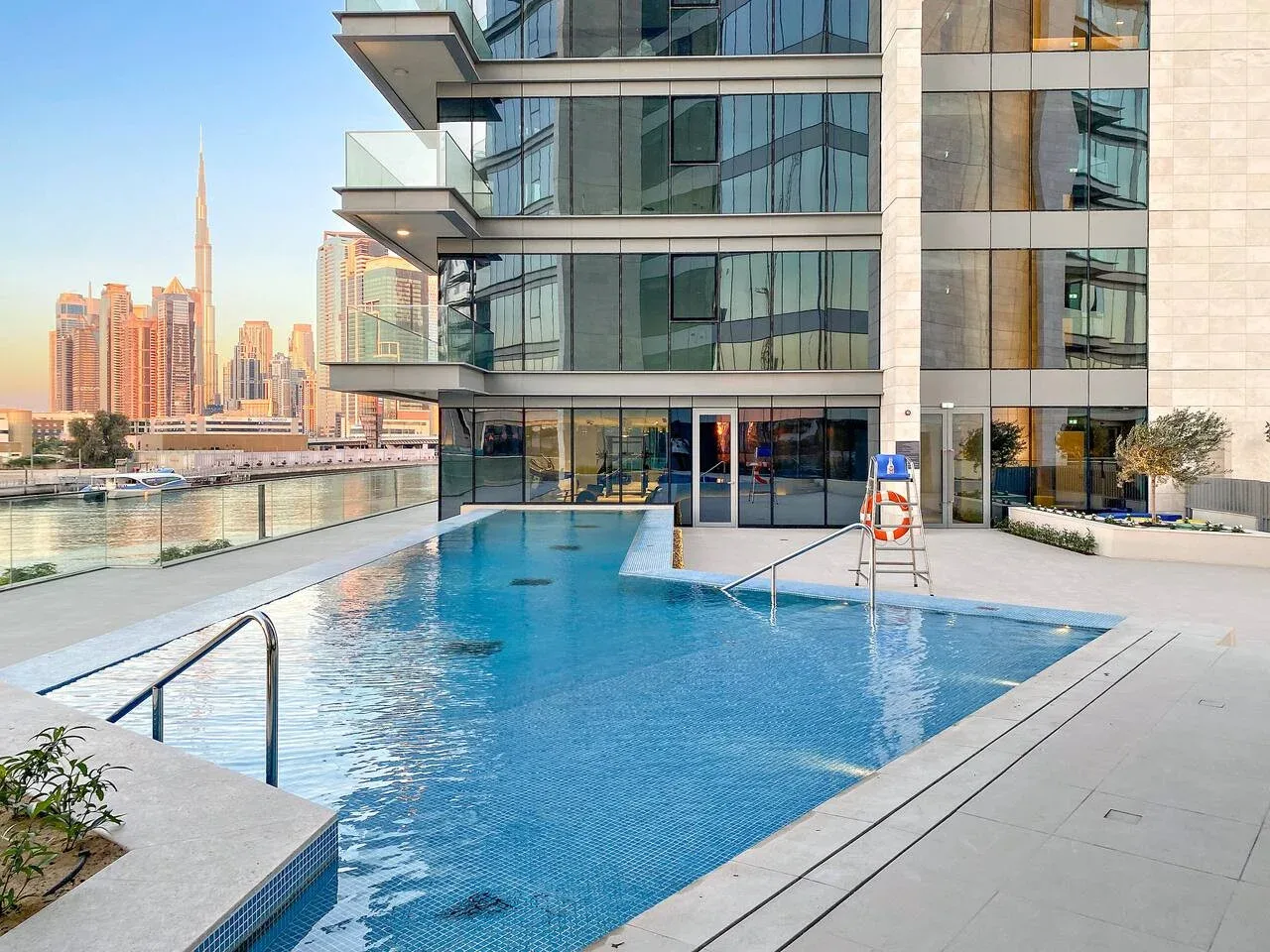 A swimming pool beside a modern glass-fronted building, with city towers in the background.