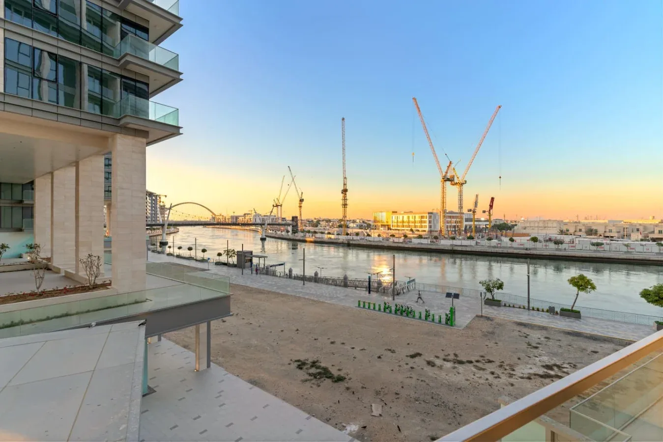 A waterfront promenade overlooks a river and construction cranes at sunset.
