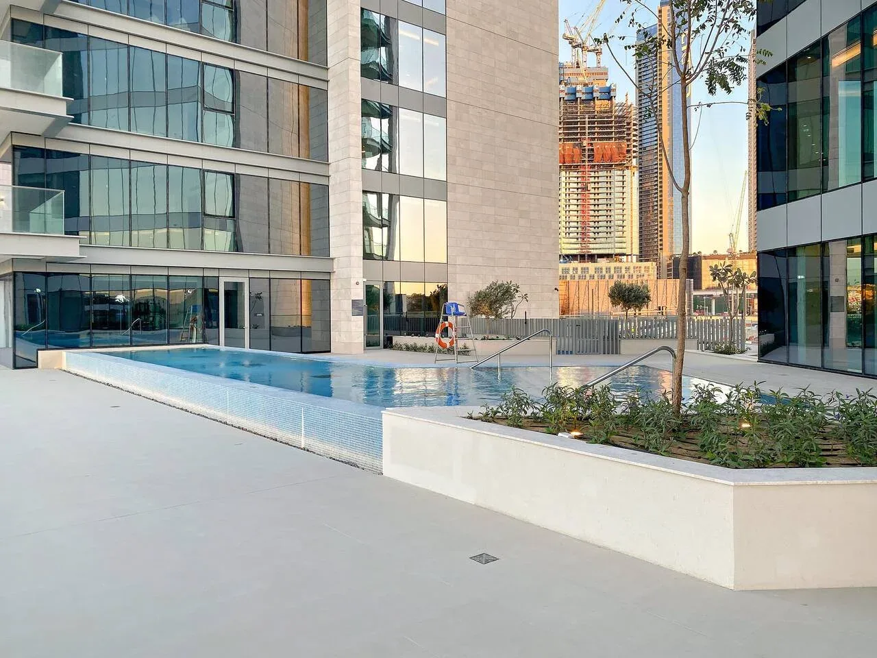 An outdoor swimming pool sits beside modern glass-fronted buildings.