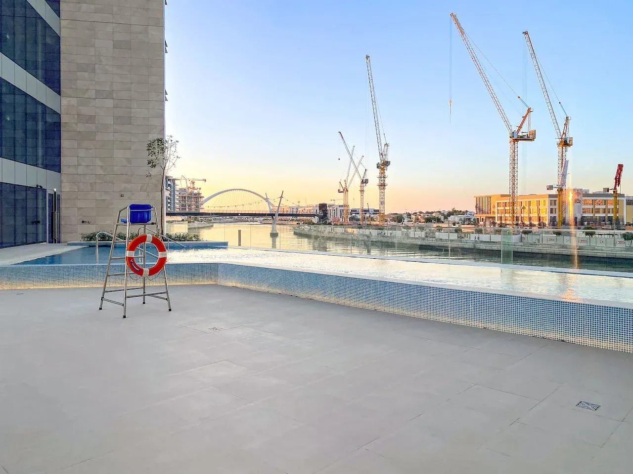 An outdoor swimming pool beside a modern building, with construction cranes visible in the distance.
