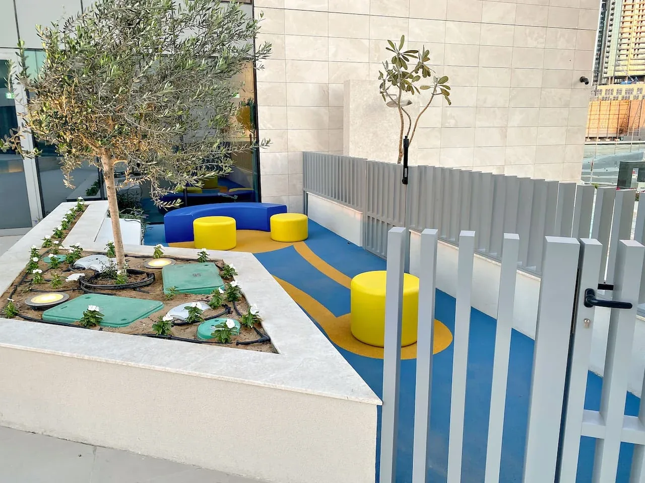 A small outdoor play area has blue flooring, yellow seating, and planted trees.