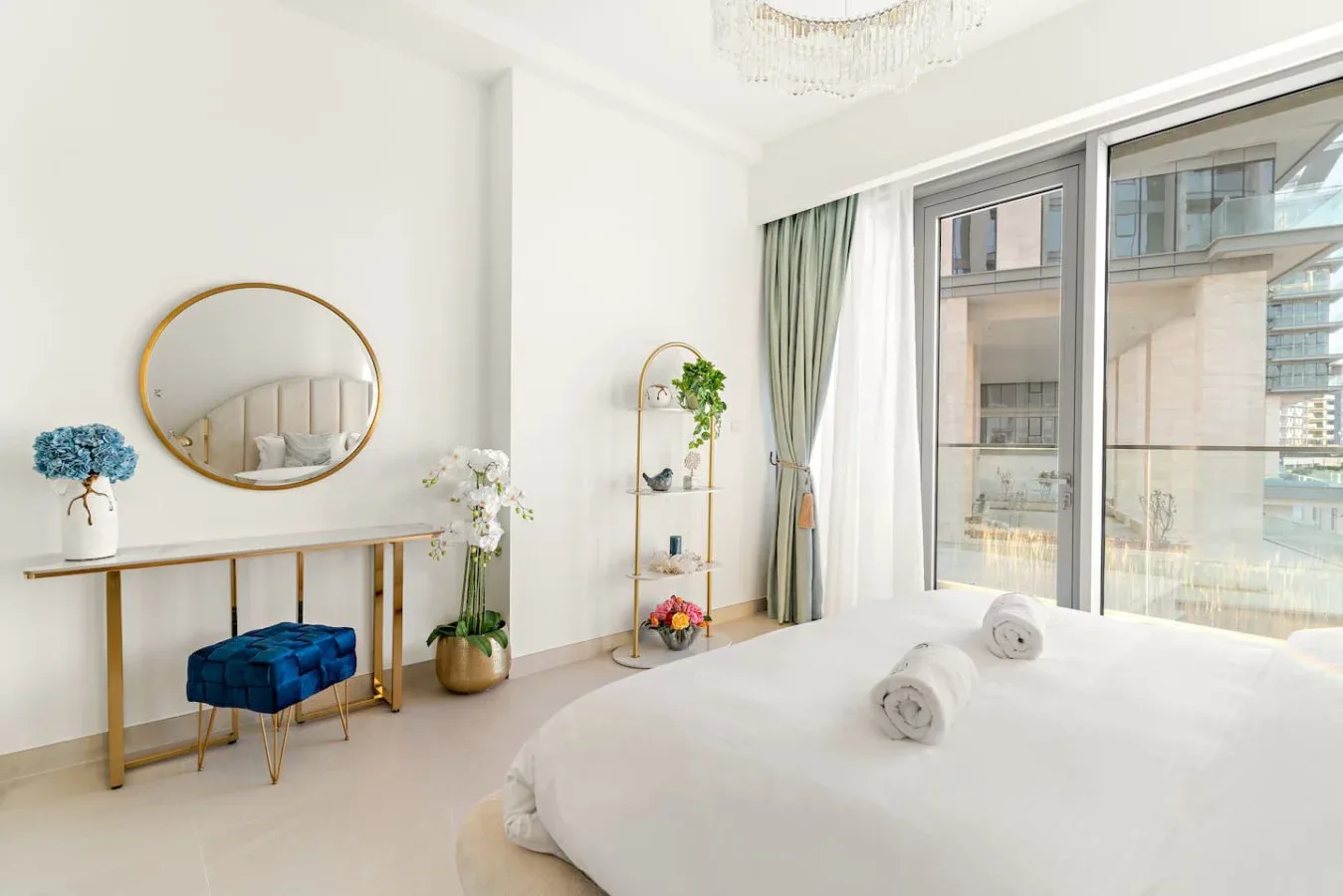 A bright bedroom with a bed, a round wall mirror, and large windows.