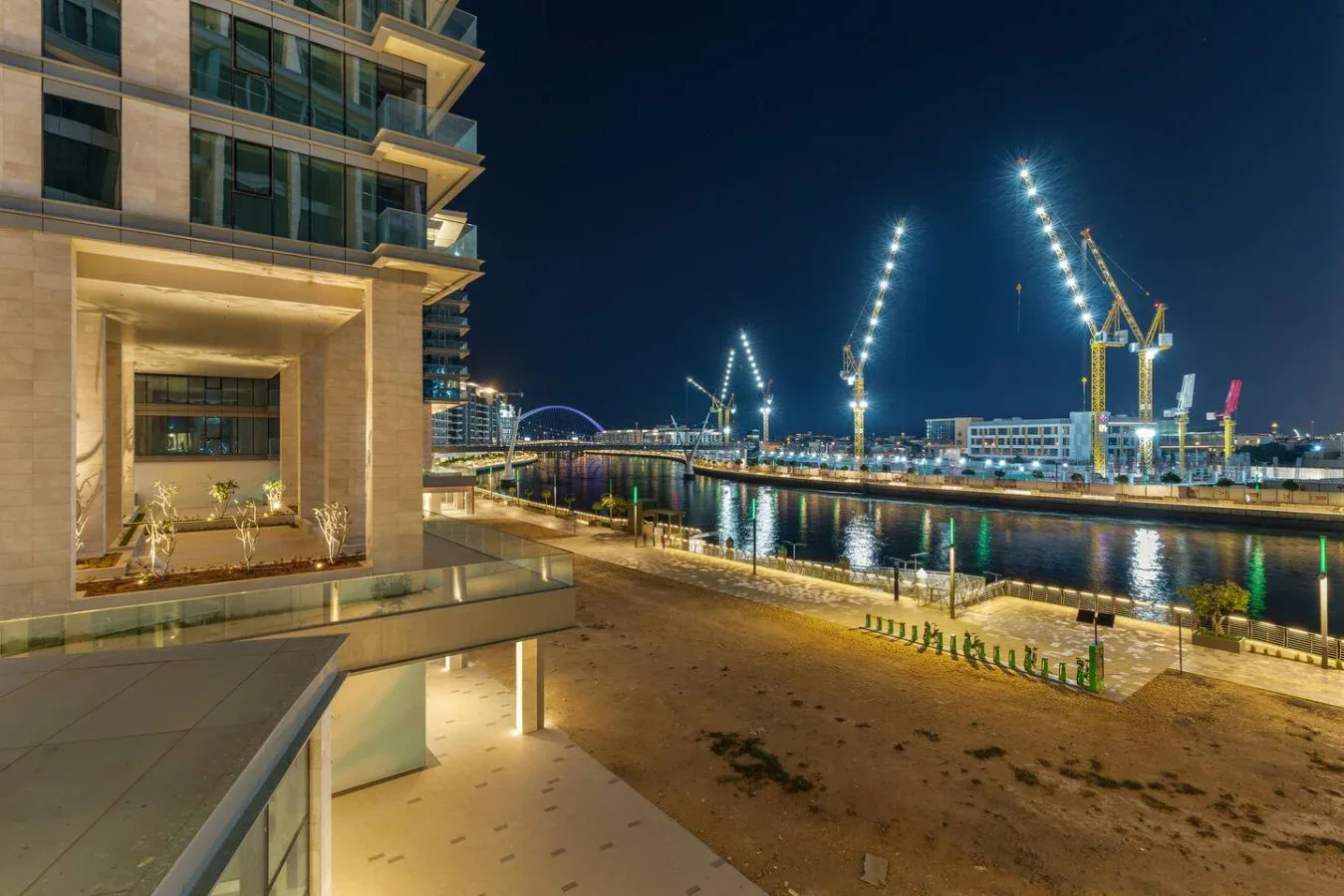 A waterfront promenade at night, with illuminated construction cranes across the water.