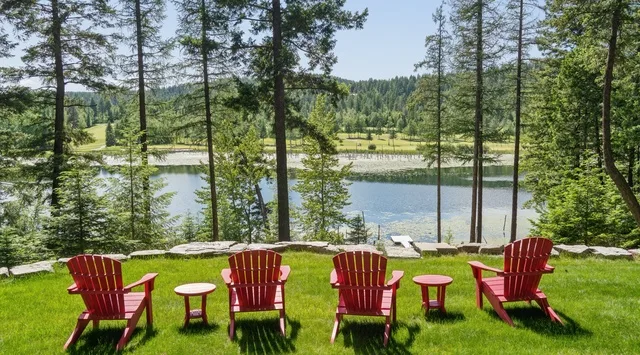 Red Adirondack chairs face a river from a grassy lawn beneath tall trees.