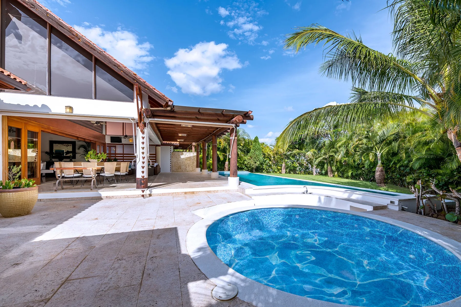 Piscina exterior junto a un patio cubierto con asientos y vegetación tropical.