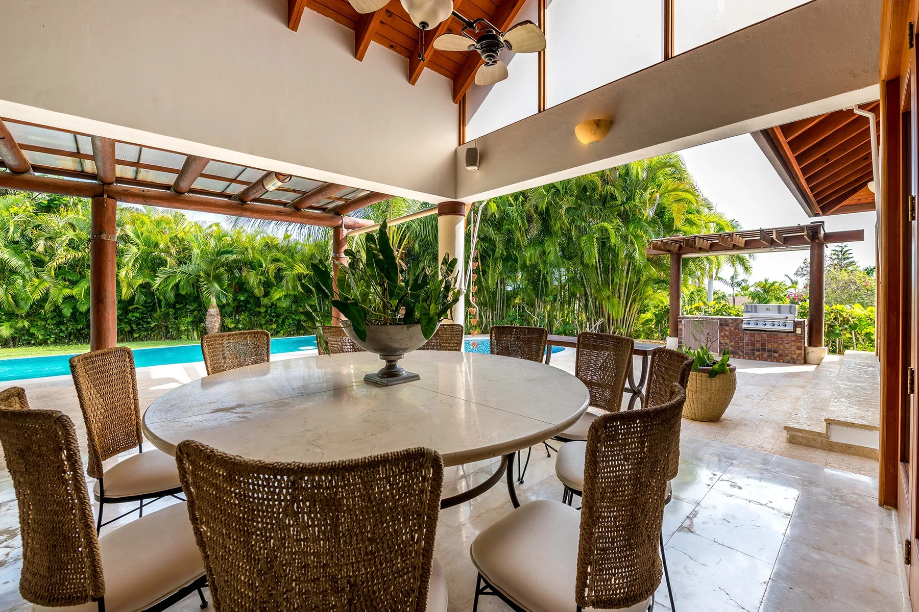 Un comedor exterior cubierto da a una piscina y una exuberante vegetación tropical.
