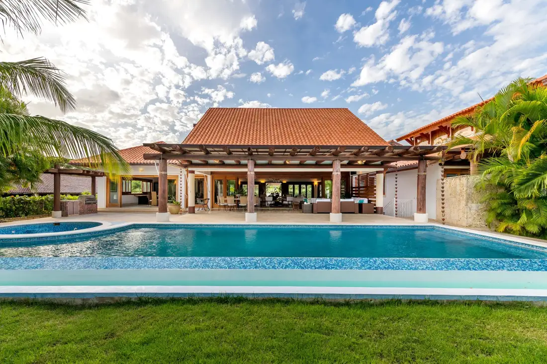 Una piscina al aire libre se encuentra frente a un patio cubierto y un jardín.
