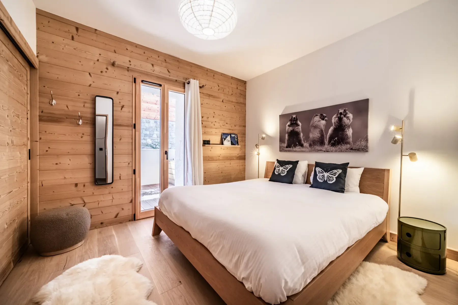 Acogedor dormitorio con paredes de madera, cama doble, almohadas con mariposas y una puerta de cristal que da a un balcón. Iluminación cálida y decoración inspirada en la naturaleza.