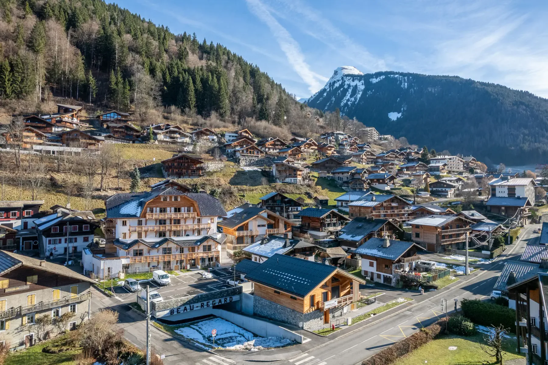 Un gran edificio de estilo chalet con balcones de madera y tejado de pizarra, rodeado de vehículos aparcados y árboles, con una ladera boscosa de fondo.