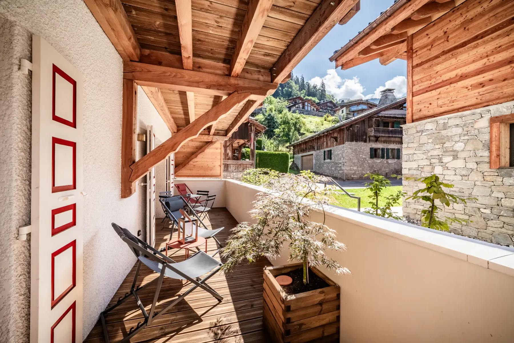 Balcón de madera con dos sillas y vistas a casas de estilo alpino y abundante vegetación bajo un cielo azul despejado.