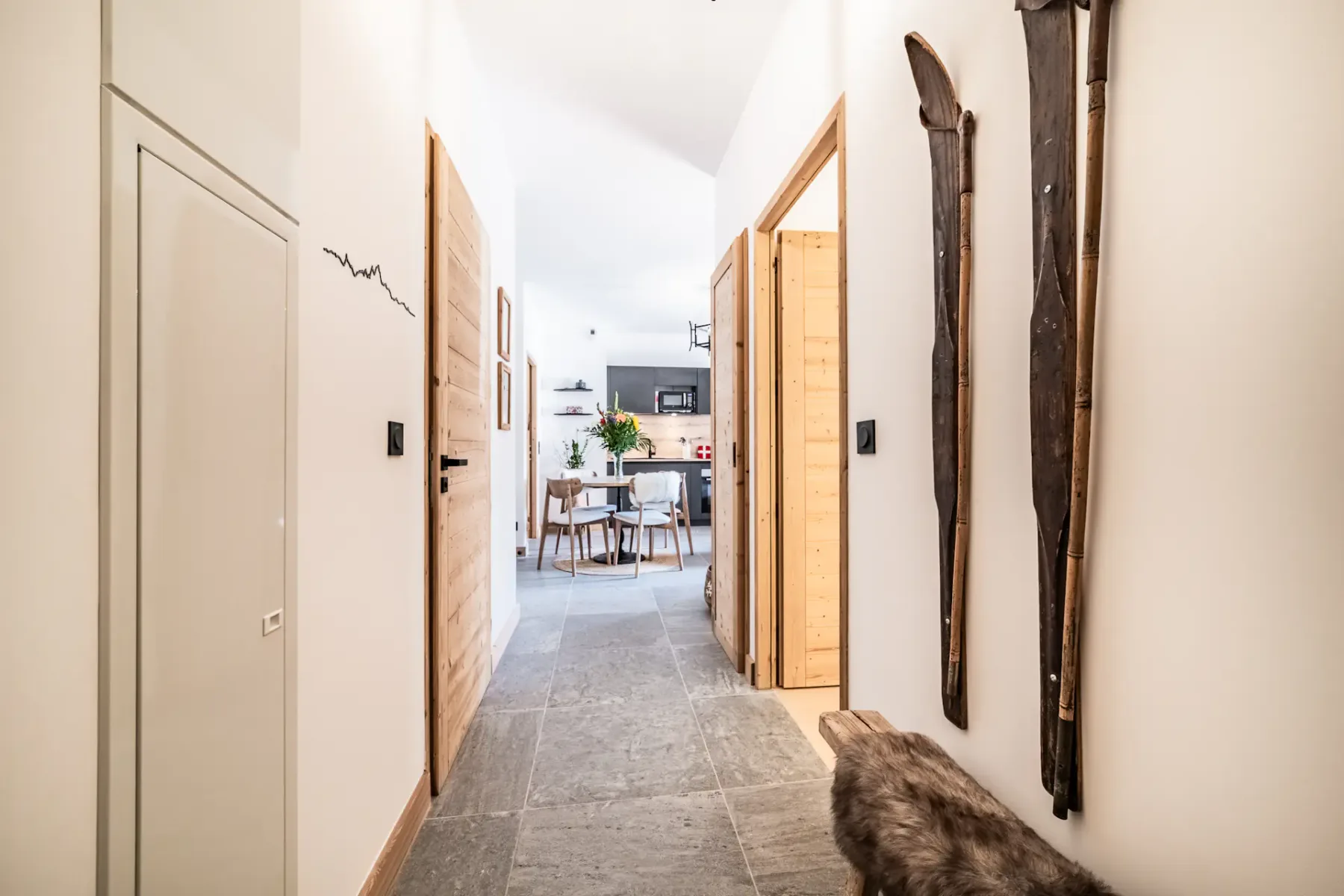 Pasillo luminoso con suelo de piedra, puertas de madera y decoración rústica, que conduce a un comedor moderno con una mesa y sillas.