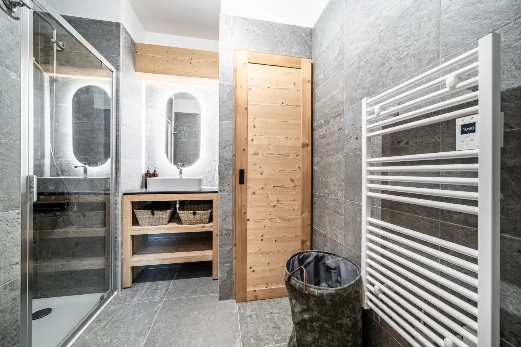 Baño moderno con azulejos grises, una puerta de madera, un mueble de lavabo con dos espejos ovalados, una ducha con mampara de cristal y un radiador toallero blanco.