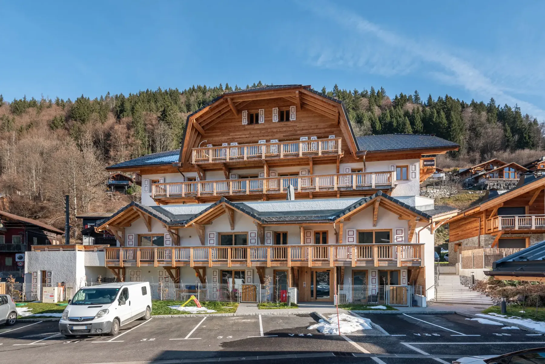 Un gran edificio de estilo chalet con balcones de madera y tejado de pizarra, rodeado de vehículos aparcados y árboles, con una ladera boscosa de fondo.