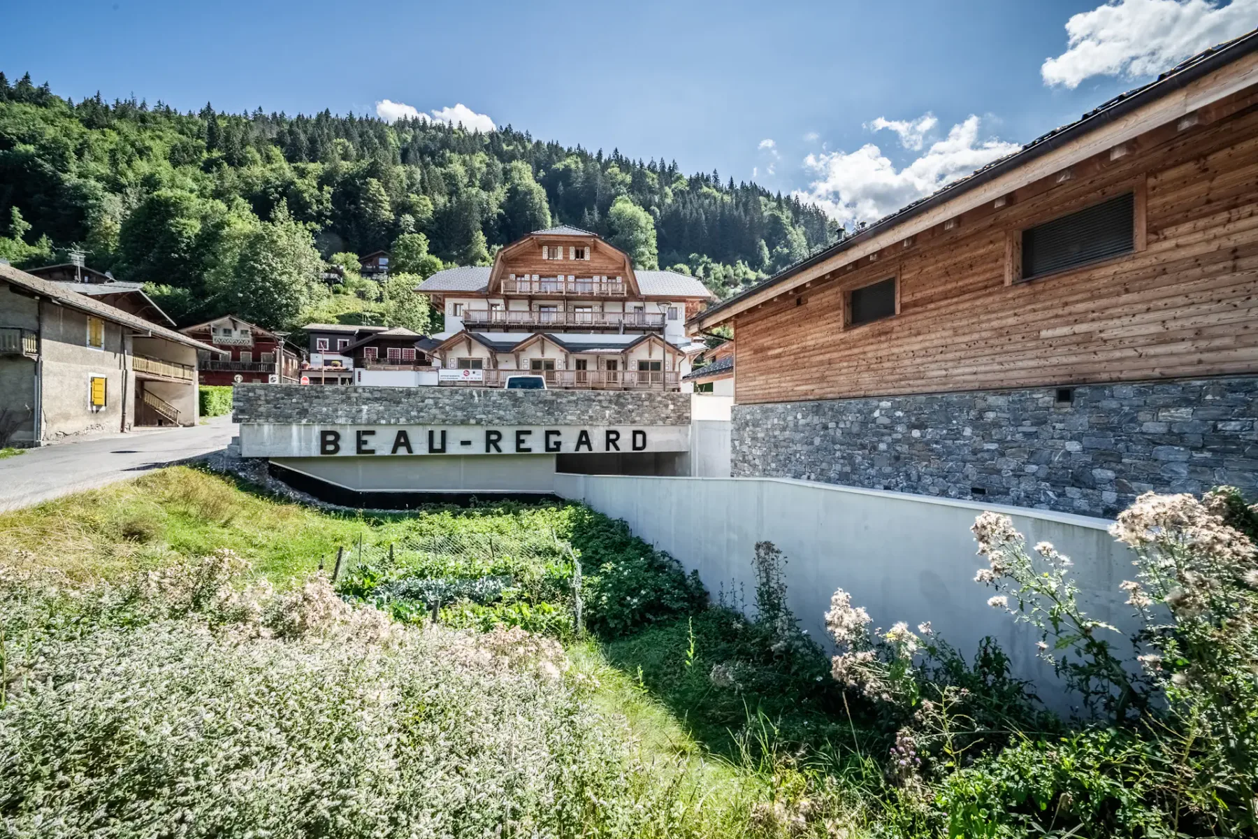 Un gran edificio de estilo chalet con balcones de madera y tejado de pizarra, rodeado de vehículos aparcados y árboles, con una ladera boscosa de fondo.