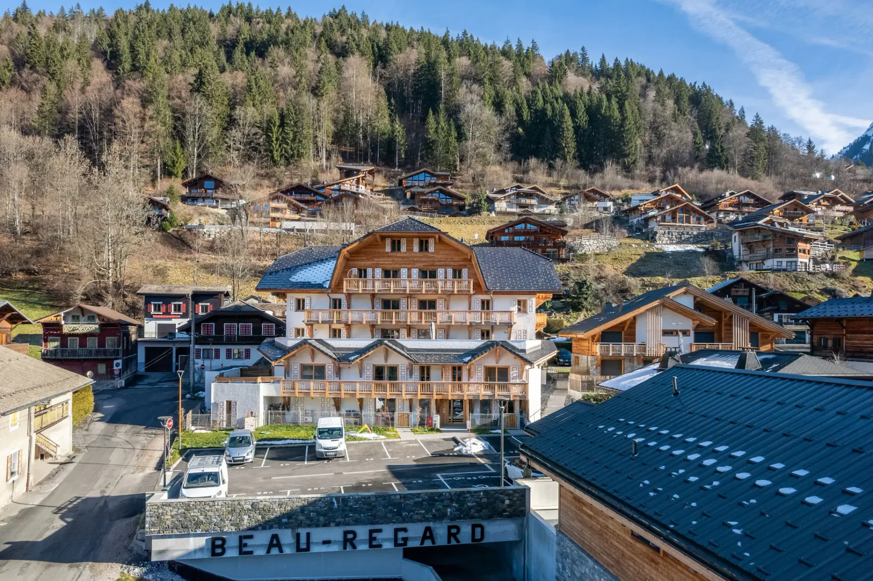 Un gran edificio de estilo chalet con balcones de madera y tejado de pizarra, rodeado de vehículos aparcados y árboles, con una ladera boscosa de fondo.
