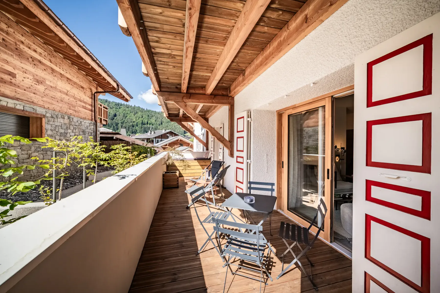 Un largo balcón de madera tiene asientos al aire libre y vistas a edificios cercanos y vegetación.