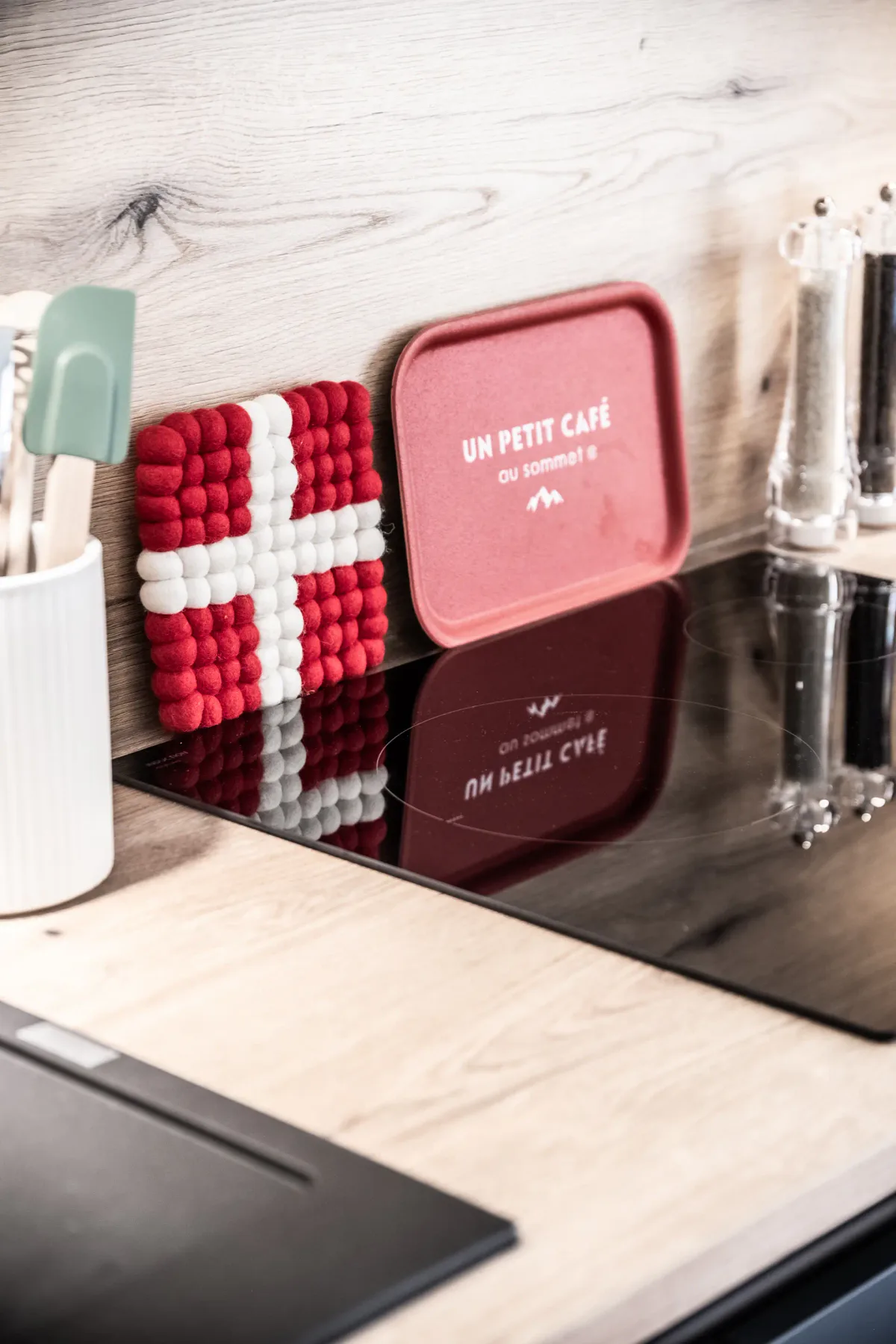 Cocina moderna con placa de inducción, posavasos con la bandera danesa, una bandeja roja con texto en francés y un recipiente para utensilios sobre una encimera de madera.