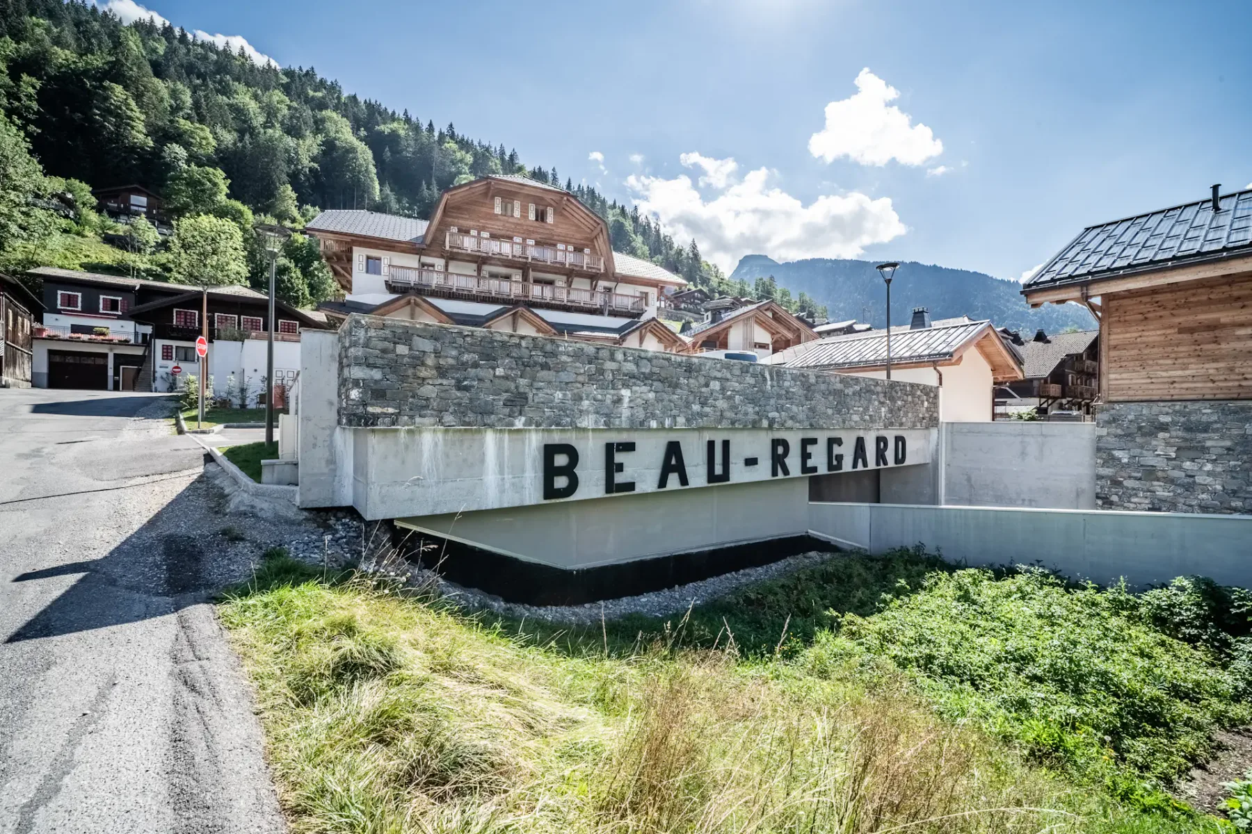 Un gran edificio de estilo chalet con balcones de madera y tejado de pizarra, rodeado de vehículos aparcados y árboles, con una ladera boscosa de fondo.