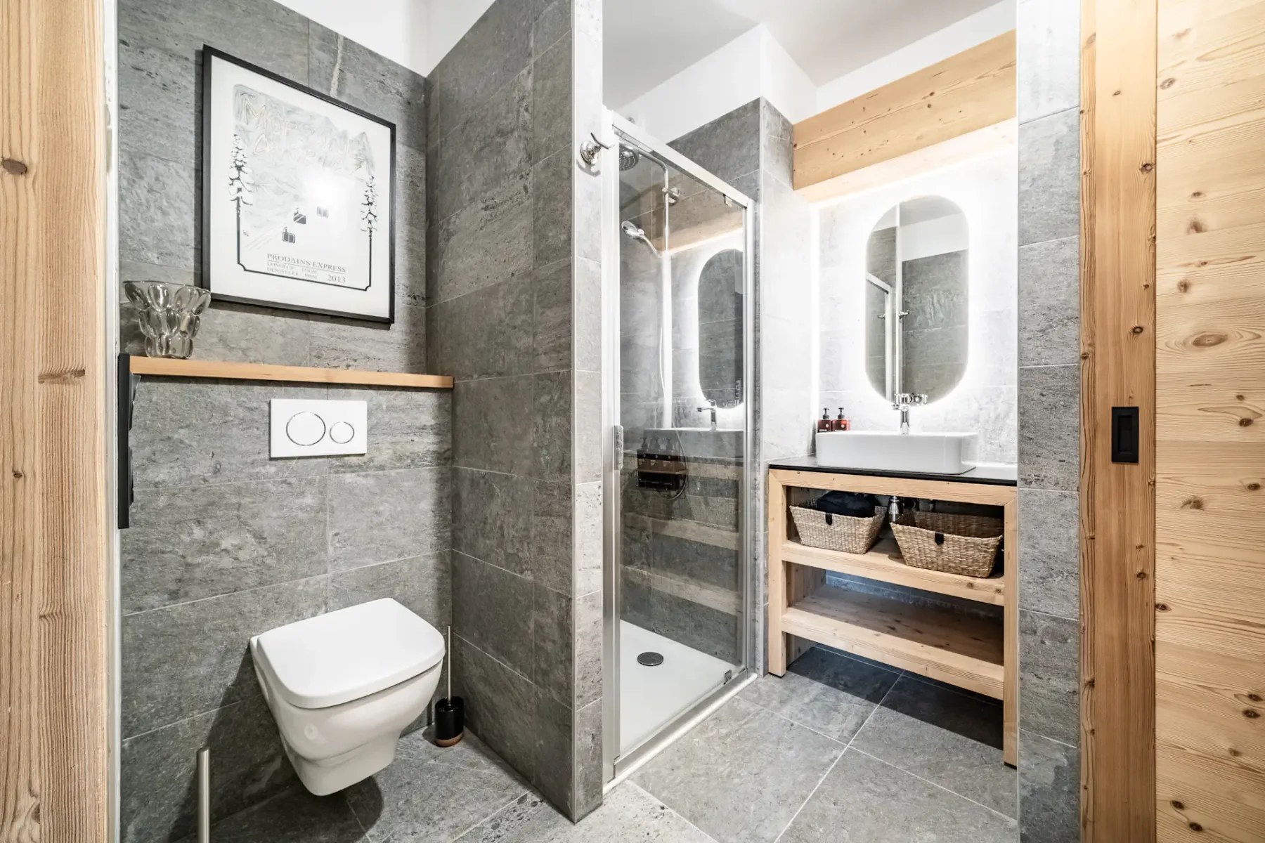 Un baño moderno con ducha de cristal y un mueble de madera bajo un espejo ovalado.