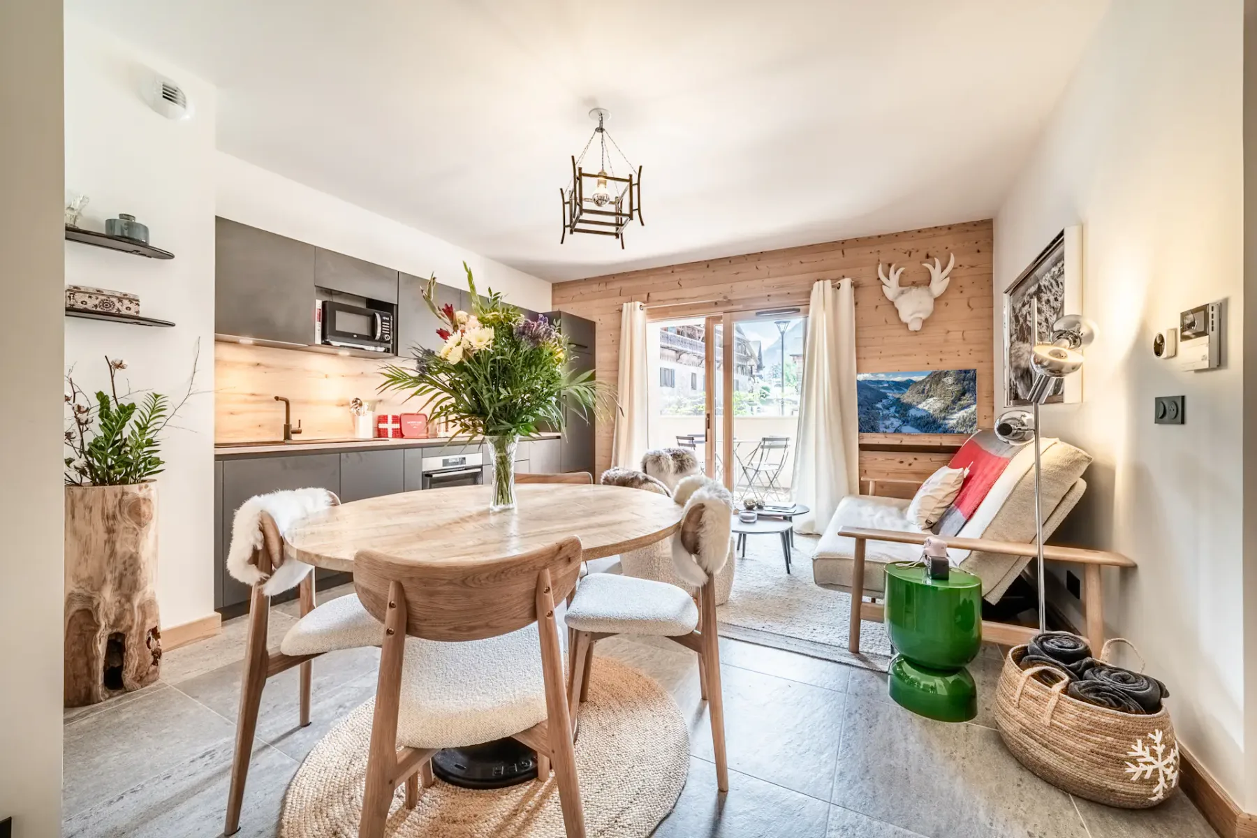 Cozy open-plan living room and kitchen with rustic decor, featuring a round dining table, modern kitchen, and seating area near a large window.