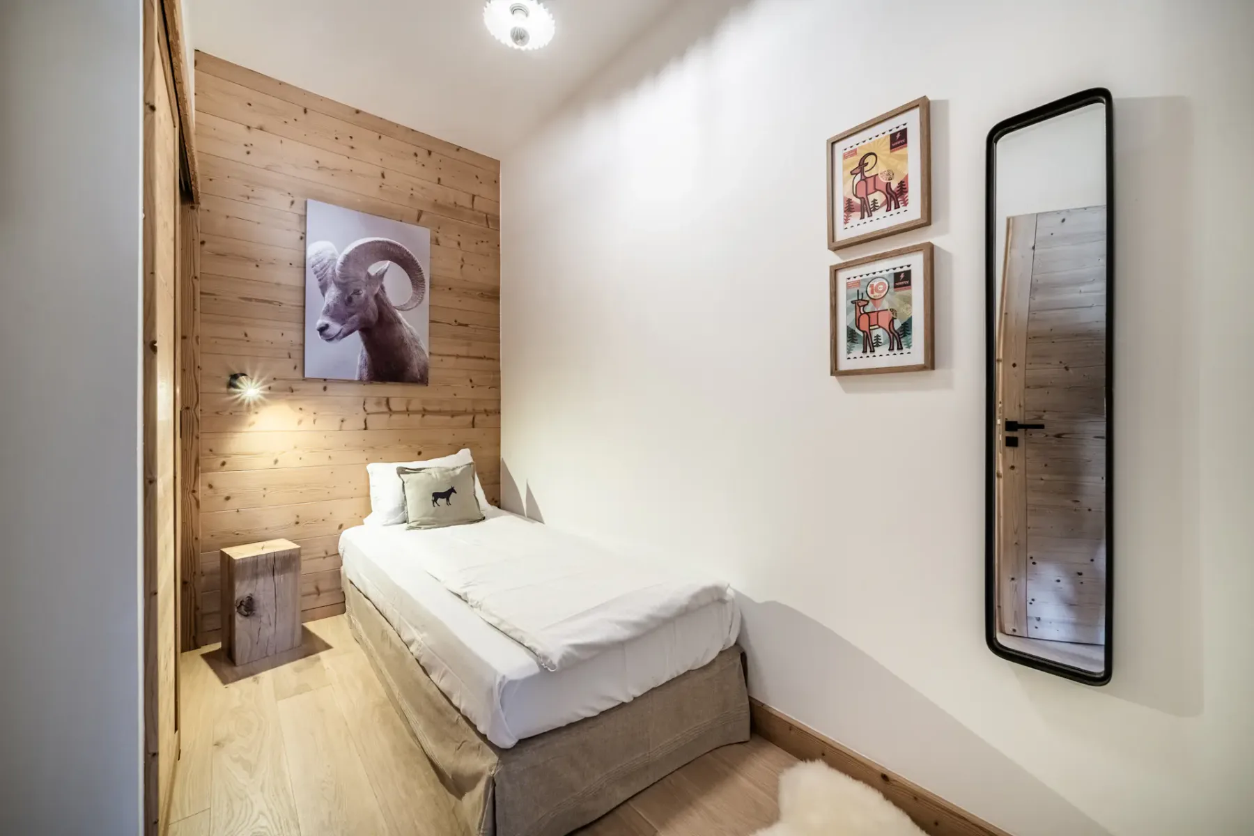 Un acogedor dormitorio con una cama individual, paredes de madera, dos obras de arte, una lámpara de pared, una mesa auxiliar y un espejo alto.
