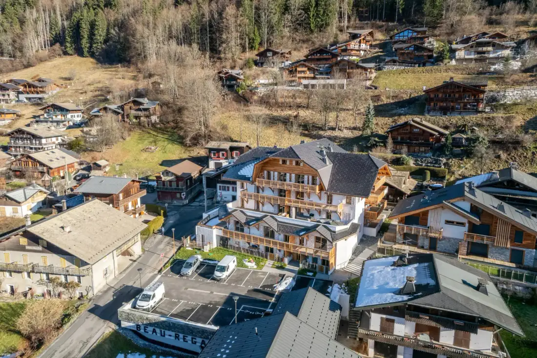 Un gran edificio de estilo chalet con balcones de madera y tejado de pizarra, rodeado de vehículos aparcados y árboles, con una ladera boscosa de fondo.