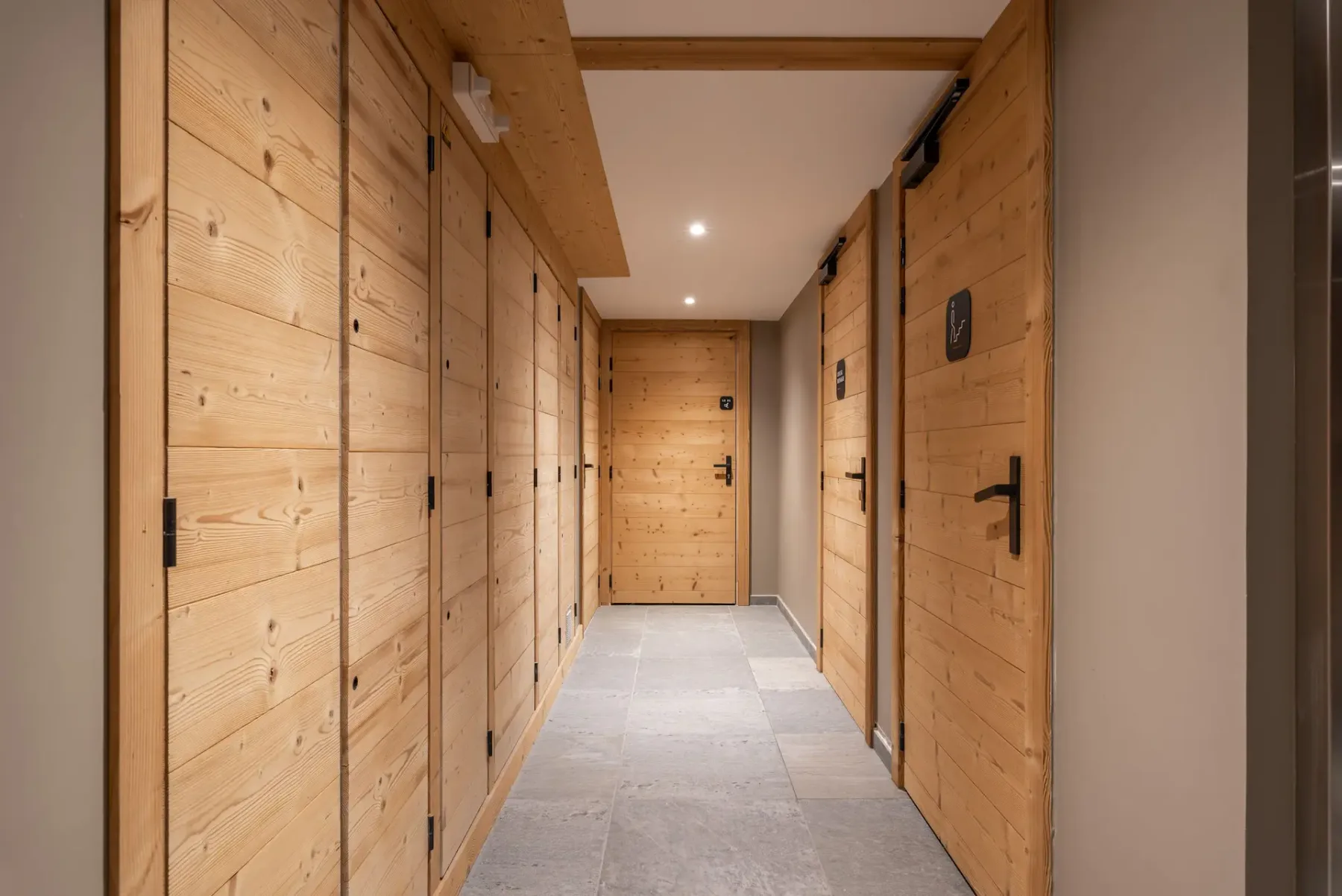 Un pasillo con puertas de madera a ambos lados y suelo de baldosas de piedra, que conduce a otra puerta de madera al fondo, iluminado por luces cálidas en el techo.
