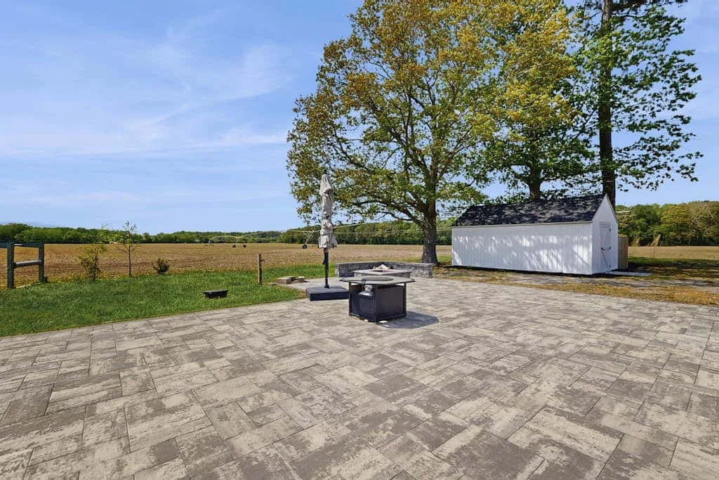 Un patio pavimentado da a un campo abierto de hierba, con una mesa y una sombrilla.