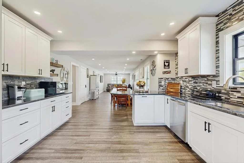 Una cocina luminosa con gabinetes blancos, encimeras oscuras y una mesa de comedor al fondo.