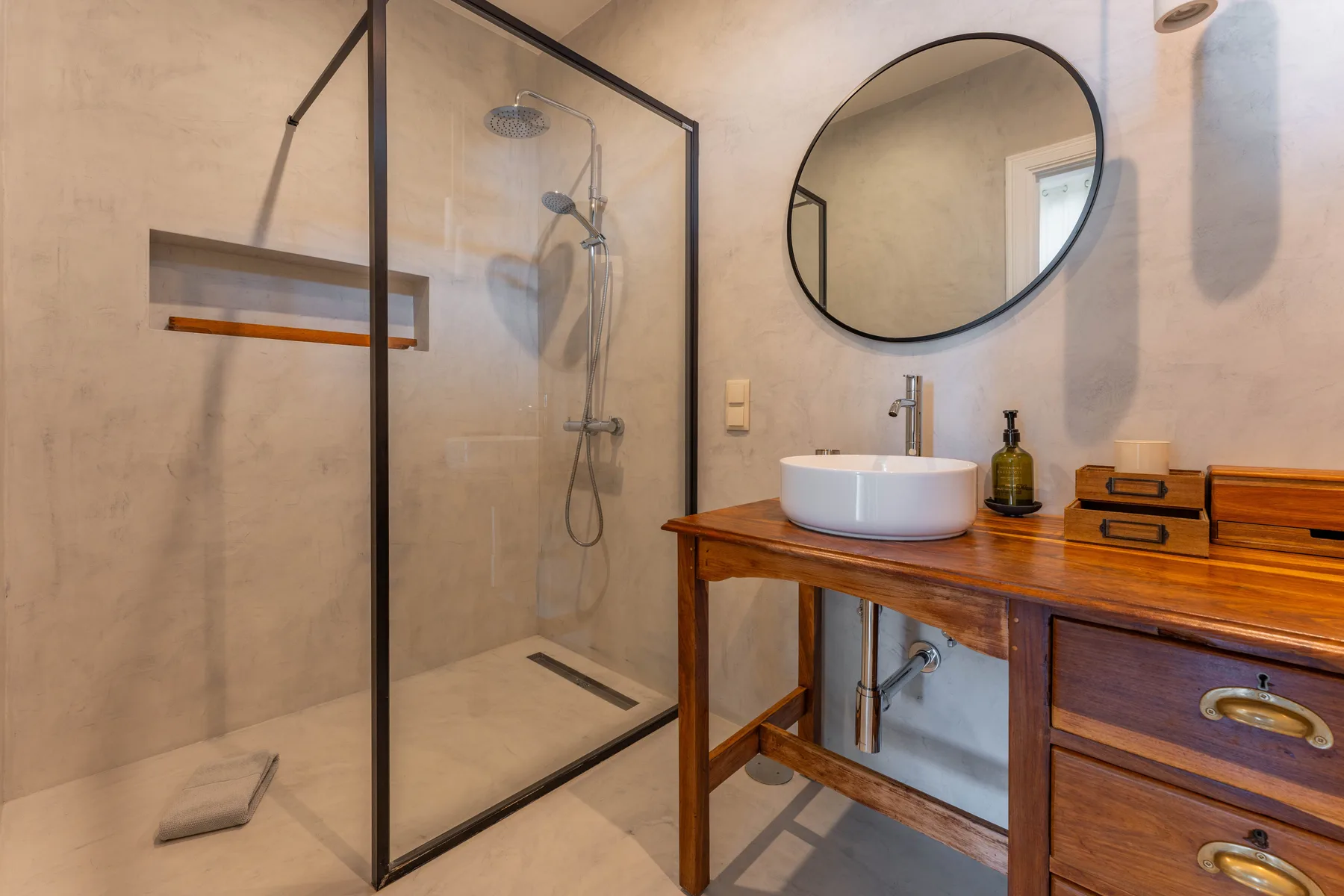 Un baño con una ducha de vidrio y un mueble de madera bajo un espejo redondo.